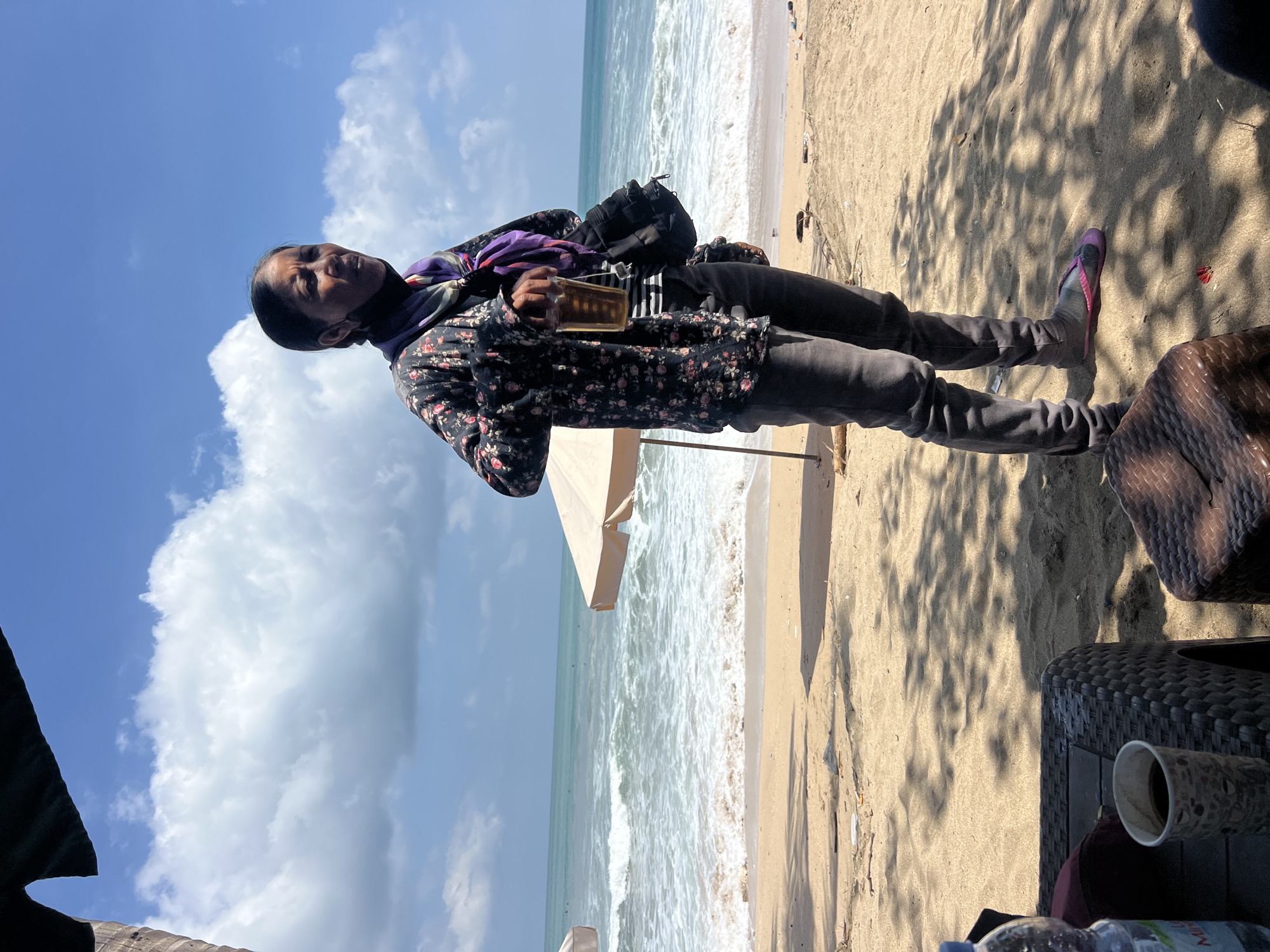 A woman stands on a sandy beach in Kuta, Indonesia, holding a glass of amber liquid. She is wearing a floral-patterned jacket, dark pants, and a purple scarf around her neck. A dark bag is slung over her shoulder. In the background, the ocean waves crash onto the shore under a partly cloudy blue sky. Beach umbrellas and some debris are visible on the sand. In the foreground, a woven table holds a bottle of water and a cup of coffee. The scene is partially shaded by trees.