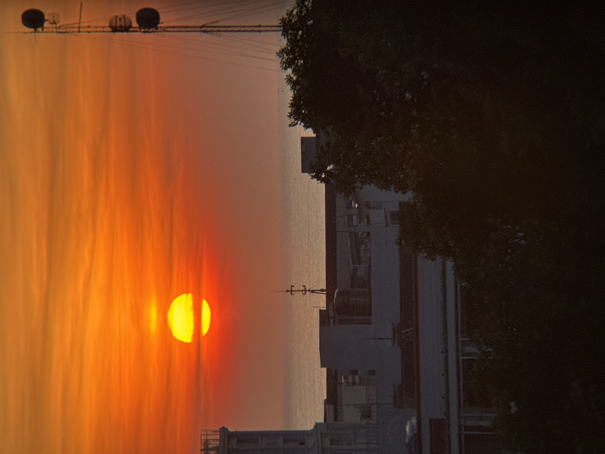 An orange sunset illuminates the sky over Nha Trang, Vietnam. The sun, a vibrant yellow orb, is partially obscured by clouds and appears low on the horizon, casting a warm glow over the scene. In the foreground, silhouetted buildings and trees create a dark contrast against the bright sky. A tall radio tower with multiple antennas stands prominently on the right side of the image, its metallic structure silhouetted against the sunset. The ocean is visible in the background, its surface reflecting the colors of the sky. The overall impression is one of a tranquil evening scene in a coastal city.