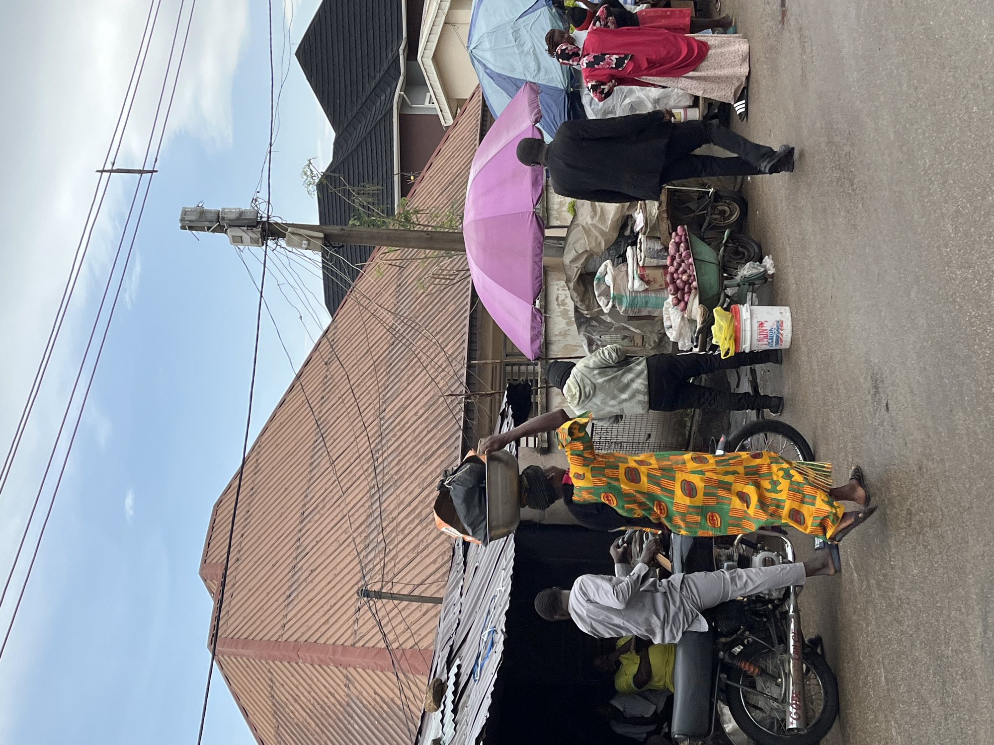 The image depicts a street scene in Jos, Nigeria. A woman in a brightly patterned yellow and green dress sits on a motorcycle. Another man in a gray shirt sits behind her on the same motorcycle. A third person, with a light green shirt, stands nearby. On the right of the street, next to the sidewalk, vendors display goods under umbrellas. The selection of goods appears to include onions, and possibly other produce or wares. The street is paved. Buildings with corrugated metal roofs line the left side of the street. A utility pole is in the background, with multiple wires extending from it. The sky is partially visible. The angle of the shot is as if it were taken at street level.