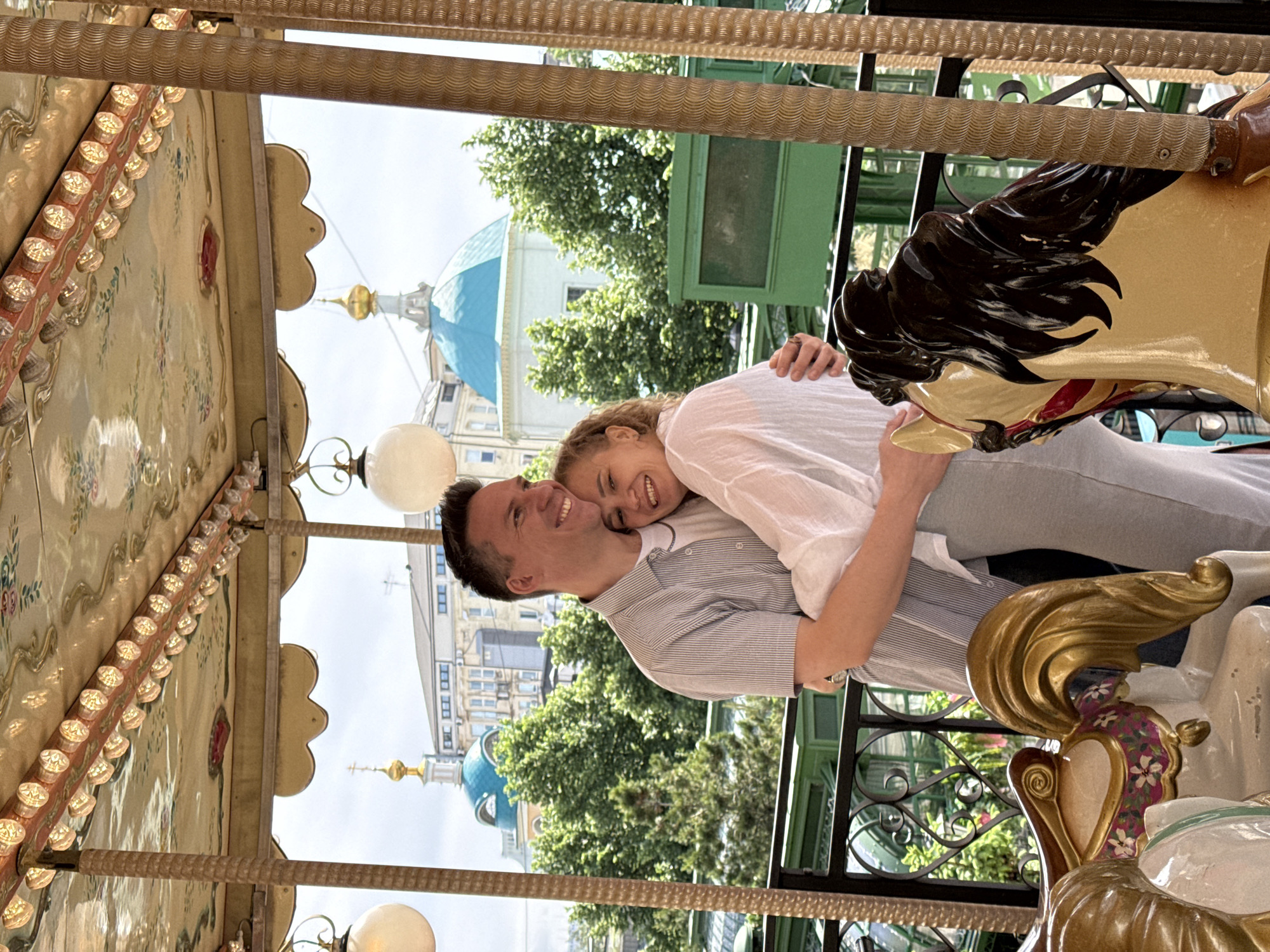 The image shows a couple on a carousel. The man is embracing the woman from behind. Both are smiling. A carousel horse is partially visible to the right of the couple. The carousel has a decorated canopy with lights. Outside the carousel, there is greenery, and in the background, a building with a distinctive blue dome and golden spire can be seen. The location is Moscow, Russia, with the building in the background being the Church of St. Nicholas on the Three Mountains.