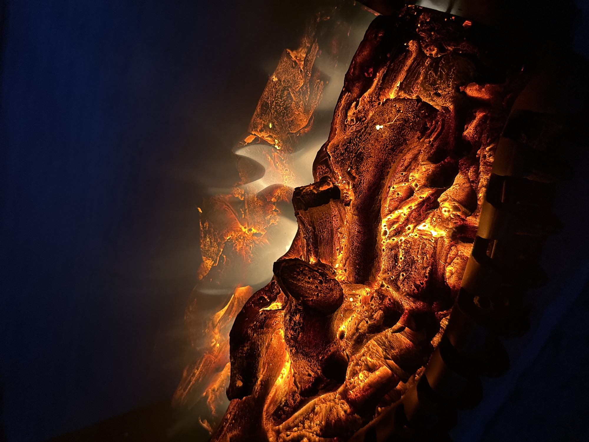 A close-up, dimly lit shot of an electric fireplace displays glowing artificial logs and flickering flames against a dark background. The logs are textured and appear to be made of ceramic or resin, with warm orange and red light emanating from within and around them. Wisps of white smoke-like material are visible, adding to the illusion of a real fire. The background transitions from a dark blue at the top to black at the bottom, with the lower portion of the fireplace's structure, which appears to be made of dark metal or wood, partially visible. There are no people or animals present in the image. The scene depicts a simulated fire, likely for decorative purposes.