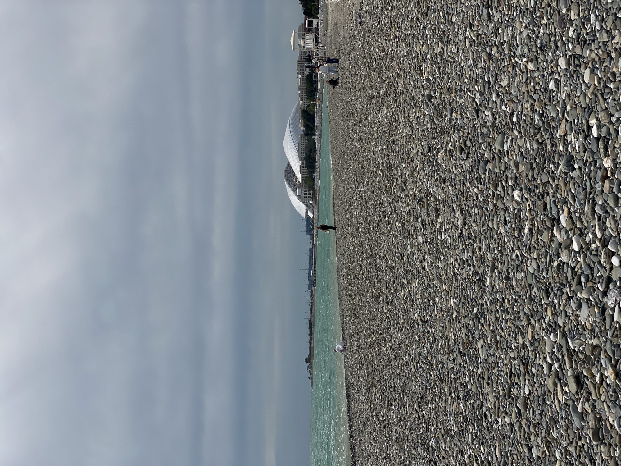 The image depicts a pebble beach on a cloudy day. In the background, the Fisht Olympic Stadium is visible along with other buildings in the city of Sirius, Russia. A few people are scattered along the beach. A person stands near the water's edge in the middle distance. Another individual is near the stadium. The water is light green, and the sky is overcast.