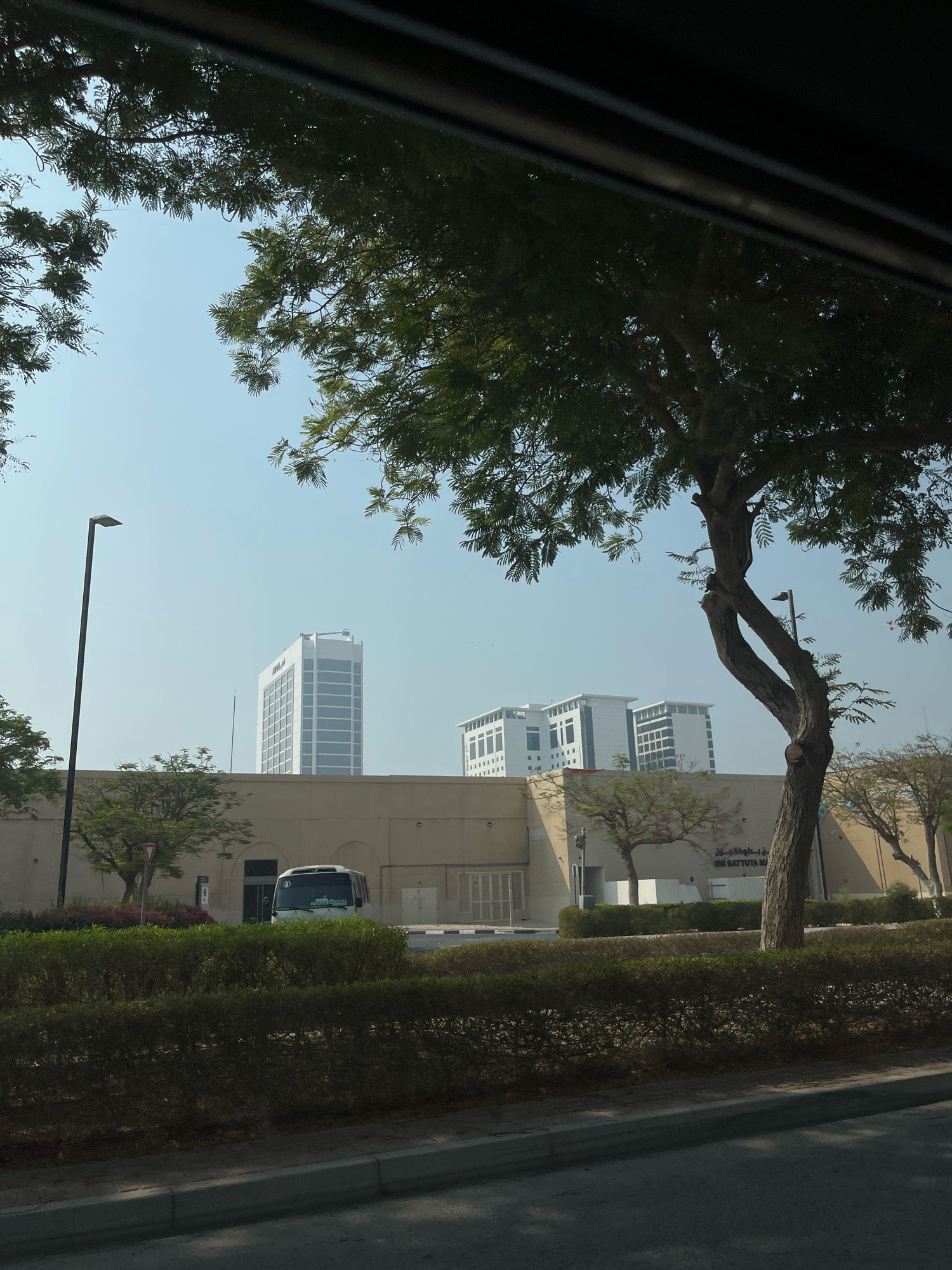 The image depicts a street view in Dubai, United Arab Emirates, likely near Ibn Battuta Mall, as indicated by the text "IBN BATTUTA MA" visible on the building. The scene is dominated by a large, mature tree in the foreground with a distinctive, gnarled trunk and branches that partially obscure the view of the background. The sky is a clear, pale blue.
In the middle ground, a long, beige building with arched entrances and windows stretches across the frame. A white bus with the number "2" displayed on its windshield is parked in front of one of the entrances, suggesting it is a mode of public transportation. Several smaller trees and shrubs are planted in front of the building, some with vibrant pink flowers.
Further in the background, two modern, multi-story buildings are visible. The one on the left is taller and predominantly white with a grid of blue-tinted windows. The building on the right is shorter and features a similar architectural style. A street lamp stands to the left of the beige building. The foreground also shows a manicured hedge and a grey curb separating the road from the sidewalk. The overall impression is an urban environment with green spaces integrated into the architecture, under a bright, sunny sky.