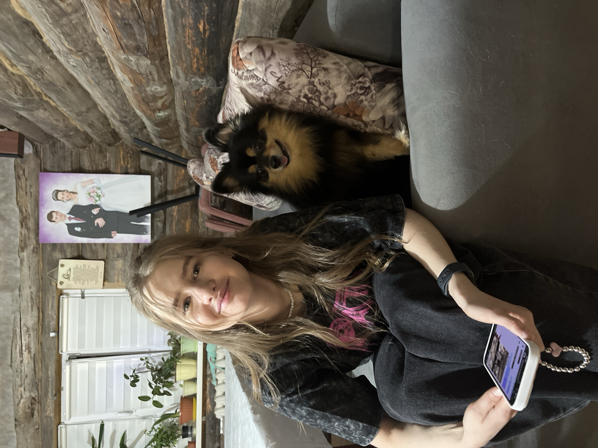 A person with fair skin and long blonde hair is seated on a gray couch, holding a white smartphone in their right hand. The person is wearing a dark gray T-shirt with a pink design and dark jeans. To the left, a small black and tan dog with its tongue slightly visible is nestled against a floral patterned pillow. Behind the couch is a wall made of horizontal wooden logs. On the left wall, above a white window with a small plant, hangs a painting depicting a wedding couple. Based on the appearance of the wall and interior design, the location appears to be inside a log cabin-style home. There is no visible evidence to indicate that this photo was taken in Izhevsk, Russia.