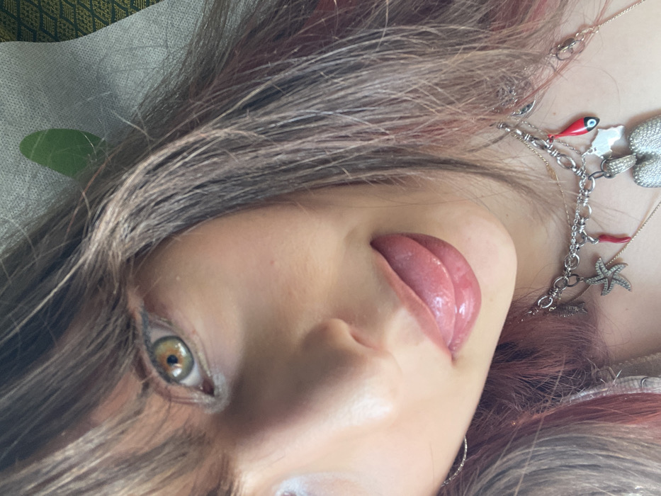An extreme close-up photograph depicts a female individual in Tzafria, Israel, positioned at an upward angle. Her left eye, characterized by a green-hazel iris with a dark pupil and eyeliner, is visible at the top center. Brown hair with reddish-pink highlights frames the left side of her face and extends across the upper forehead as bangs. Her mouth is partially open, revealing a glossy, pink-toned tongue extended outwards. Multiple layered necklaces are visible below her chin, including a silver-toned choker adorned with various charms: a starfish, a red chili pepper, a fish, a white outline charm, and a large textured heart-shaped pendant. A subtle silver hoop earring is present on the left side of her face. The background consists of a light-colored, textured surface on the upper right, partially obscuring a dark green, patterned fabric.
