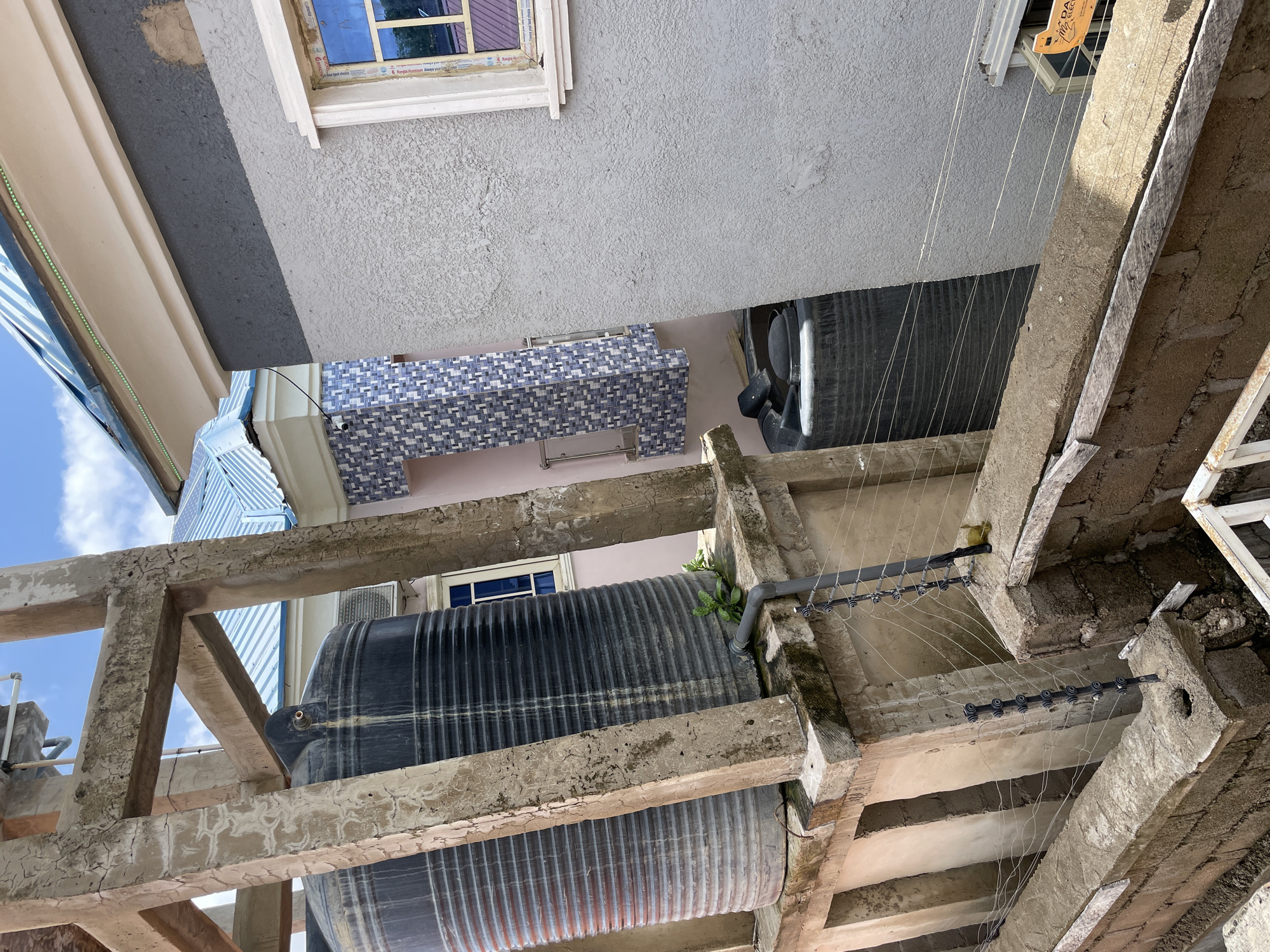This image captures an outdoor urban residential scene in Jalingo, Nigeria, viewed from a slightly elevated perspective. The central focus is a weathered concrete support structure, exhibiting visible cracks and flaking, which elevates two large, black, corrugated plastic water tanks. A grey PVC pipe connects to the base of the left tank, where a small green plant is growing, indicating past or present water overflow.
To the immediate right of the tanks, a building with a light grey textured stucco wall is visible. This wall transitions to a darker grey band, then a lighter beige section beneath the roofline. A window with a white frame and blue-tinted reflective glass is set into the light grey wall. A thin green LED strip light is affixed under the eaves of this building.
In the background, another building features a light blue corrugated metal roof. Below it, a section of wall is clad with blue and white patterned square tiles. A small window with a blue frame is also discernible on this structure. The sky above is clear blue with sparse white clouds.
In the foreground and lower portion of the image, a concrete block wall acts as a barrier or fence. Mounted on this wall is a multi-strand electric fence system, characterized by black vertical posts with numerous white ceramic or plastic insulators holding thin horizontal wires. A yellow object rests on the right side of this fence structure. Below the main tanks, a third black water tank is partially visible at ground level. A partially visible orange and white sticker, displaying "DAWA ELECTR," is affixed near the window on the right-hand building. A white metal railing is partially seen in the bottom right corner. No individuals are present in the scene.