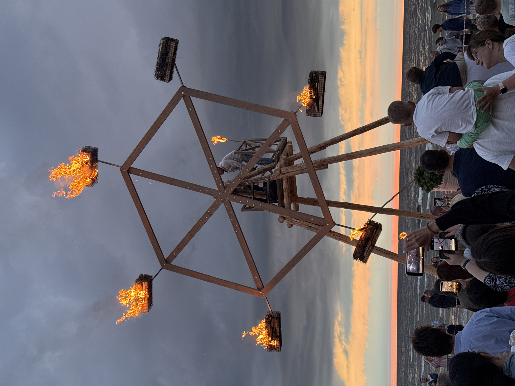 This is a medium shot photograph of a fire art installation resembling a Ferris wheel on a beach at dusk, likely in Jūrmala, Latvia, given the context of the prompt. The structure, made of wooden beams, has six arms, each with a basket at the end containing burning logs. Flames erupt from these baskets, with one at the top of the structure being the largest. A person is visible on a platform within the central structure of the installation, holding a flaming torch. In the foreground, a crowd of people are gathered, many holding up smartphones to capture the event. The background shows the Baltic Sea with a colorful sunset sky above it, featuring streaks of orange and pink clouds against a dark grey sky. The overall atmosphere suggests an outdoor performance or festival.