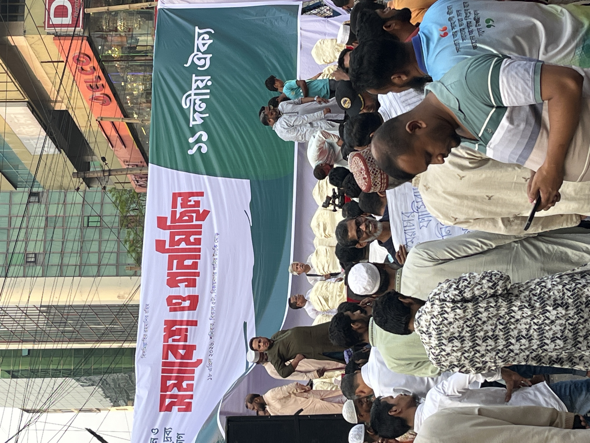 A street-level, medium-distance shot captures a political gathering in Dhaka, Bangladesh. A large banner displays text in Bengali, including "১১ দলীয় ঐক্য" (11-Party Unity) and "সমাবেশ ও প্রান মিছিল" (Rally and Procession). It also mentions the date "১৮ এপ্রিল ২০২৬" (April 18, 2026), the day "শনিবার" (Saturday), the time "বিকাল ৫টা" (5 PM), and the location "বিজয় নগর পানির টাংকি মোড়" (Bijoy Nagar Water Tank Intersection). The foreground is filled with attendees, primarily men wearing various traditional and casual clothing. Several individuals are wearing white prayer caps. In the background, buildings with commercial signs are visible, including "JELCO" and "D". Power lines crisscross the sky. The perspective is slightly angled, with the camera positioned somewhat low to the ground.