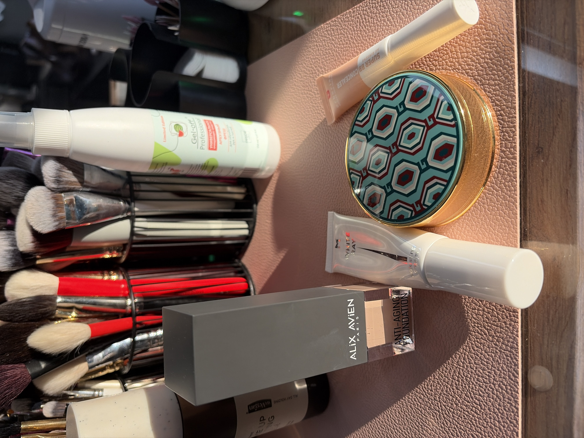 This image depicts a collection of makeup items arranged on a textured surface, possibly a makeup drawer or storage compartment. The products include a white spray bottle labeled "Gel-off Professional," a tube of "SUPER BB CONCEALER," a round container with a patterned lid, a tube labeled "WHITE RAY," and a gray box of "ALIX AVIEN PARIS ANTI-AGING FOUNDATION." The items are partially obscured by a container of makeup brushes with red and silver handles. Some text on the products is in Russian. The location is not evident from the image.