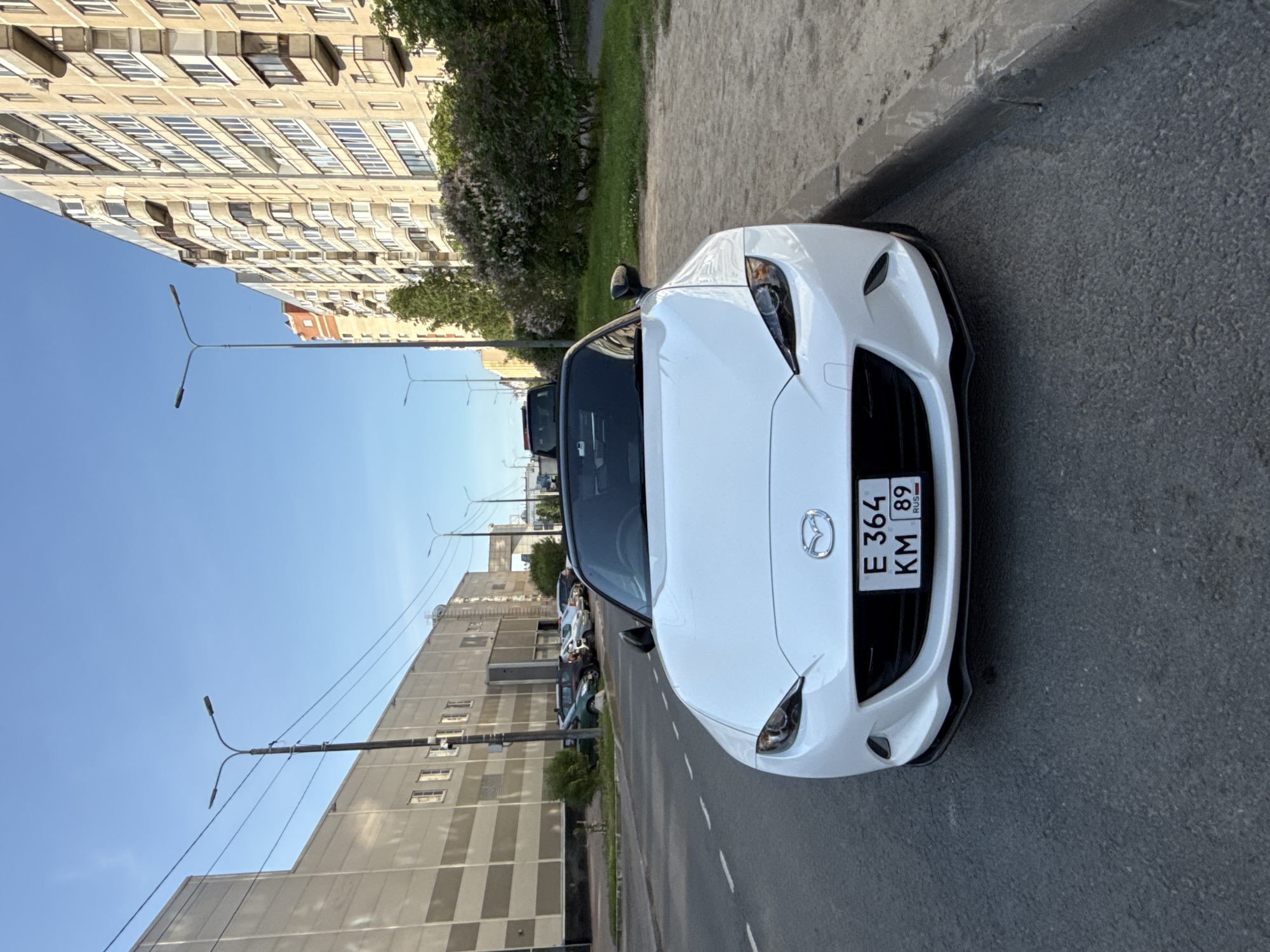 A white Mazda MX-5 convertible is parked on the side of a street in a city, likely Saint Petersburg, Russia, based on the license plate format. The car is facing the camera, with its front bumper and grille clearly visible. The license plate reads "E 364 KM 89 RUS". The street is paved, with a sidewalk to the right of the car, made of sand and gravel, and a curb separating the street from the sidewalk. In the background, there are apartment buildings, a large commercial building, streetlights, and trees with green foliage and some purple flowering bushes. The sky is clear and blue. Several other cars are parked along the street in the background. The image appears to be taken during daylight.
