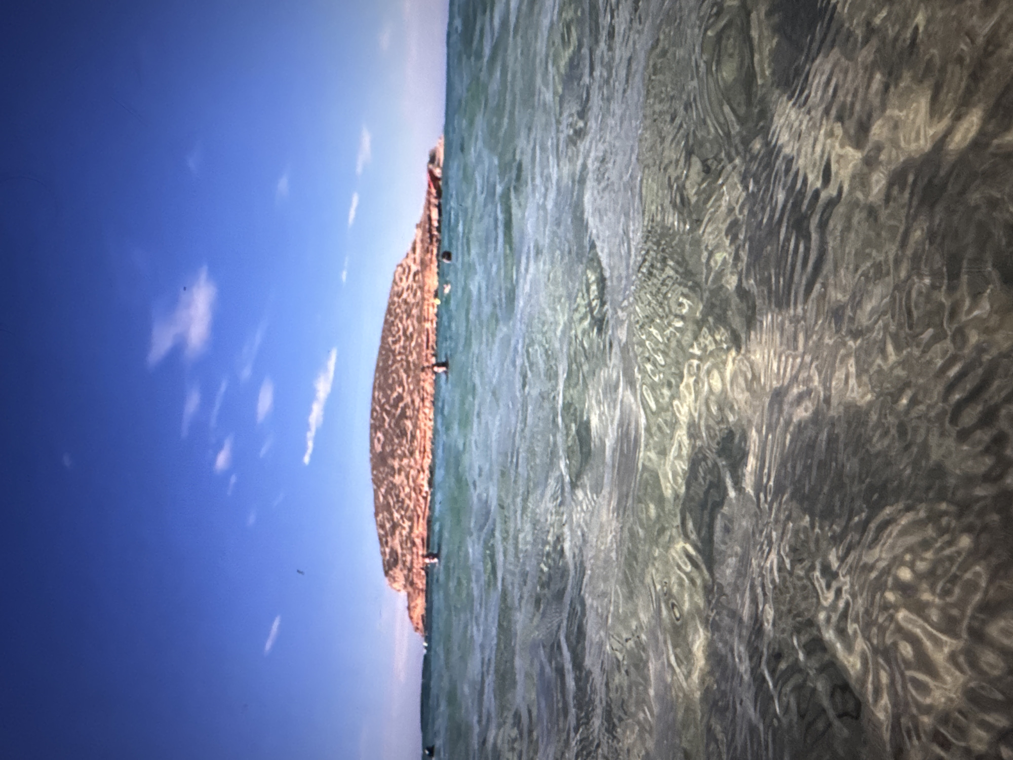 The image shows a vast expanse of clear, turquoise water in the foreground, with ripples and sunlight creating a dynamic pattern on the surface. In the mid-ground, several people are visible in the water, some appearing to be swimming or wading. Further in the distance, a large, rocky island or promontory dominates the horizon. This landmass is covered in reddish-brown vegetation and appears to be a prominent natural feature. The sky above is a vibrant blue, with a few wispy white clouds scattered across it. The overall scene depicts a lively beach or coastal area, with people enjoying recreational activities in a natural setting. The image does not contain any elements that would allow for identification of the specific location within the city of Porrentruy, Switzerland.