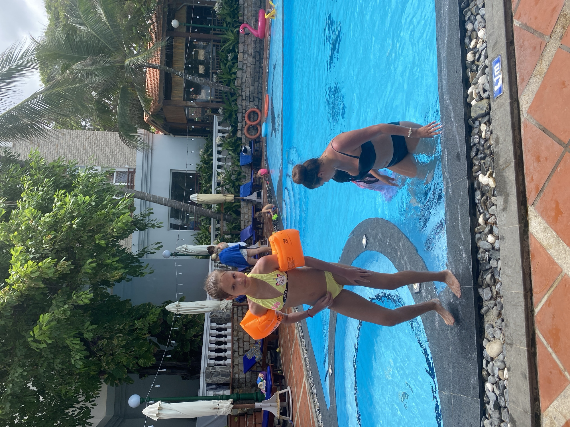 In this vibrant, sun-drenched image taken in Mũi Né, Vietnam, a young girl sporting orange arm floaties and a yellow bikini stands at the edge of a bright blue swimming pool. Her expression is one of calm engagement with the camera. To her right, a woman in a black bikini is partially submerged in the water, her attention directed towards the young girl. The pool area is bordered by a stone pathway and terra cotta tiles. In the background, lush green foliage and palm trees suggest a tropical setting. Behind the trees, a building with a terracotta roof and white walls can be seen, hinting at a resort or hotel. Other individuals are visible lounging on chairs or walking around the pool area, adding to the sense of a relaxed, holiday atmosphere. The bright sunlight and clear blue water indicate it's a sunny day, likely late morning or afternoon, perfect for a swim.