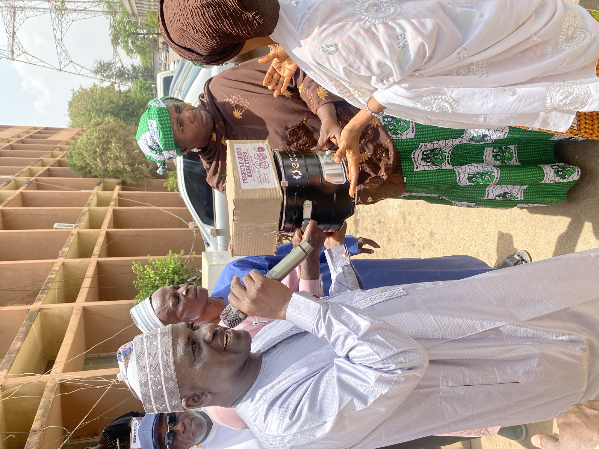 In Unguwar Rimi, Nigeria, a group of people are gathered outdoors near a light brown building. A man in a light purple traditional outfit holds a microphone and speaks. He is surrounded by other men wearing traditional Muslim caps, one of whom wears sunglasses and a blue cap. Two women stand to the side, one wearing a brown dress and a brown headscarf and the other wearing a white outfit with green and white patterned fabric. They appear to be examining a black device labeled "eco" with a box labeled "PRESTINE GREEN BRIQUETTES" attached. The fabric of the woman's dress includes text: "NATIONAL COUNCIL FOR WOMEN SOCIETIES, NIGERIA". A white vehicle is parked behind them.
