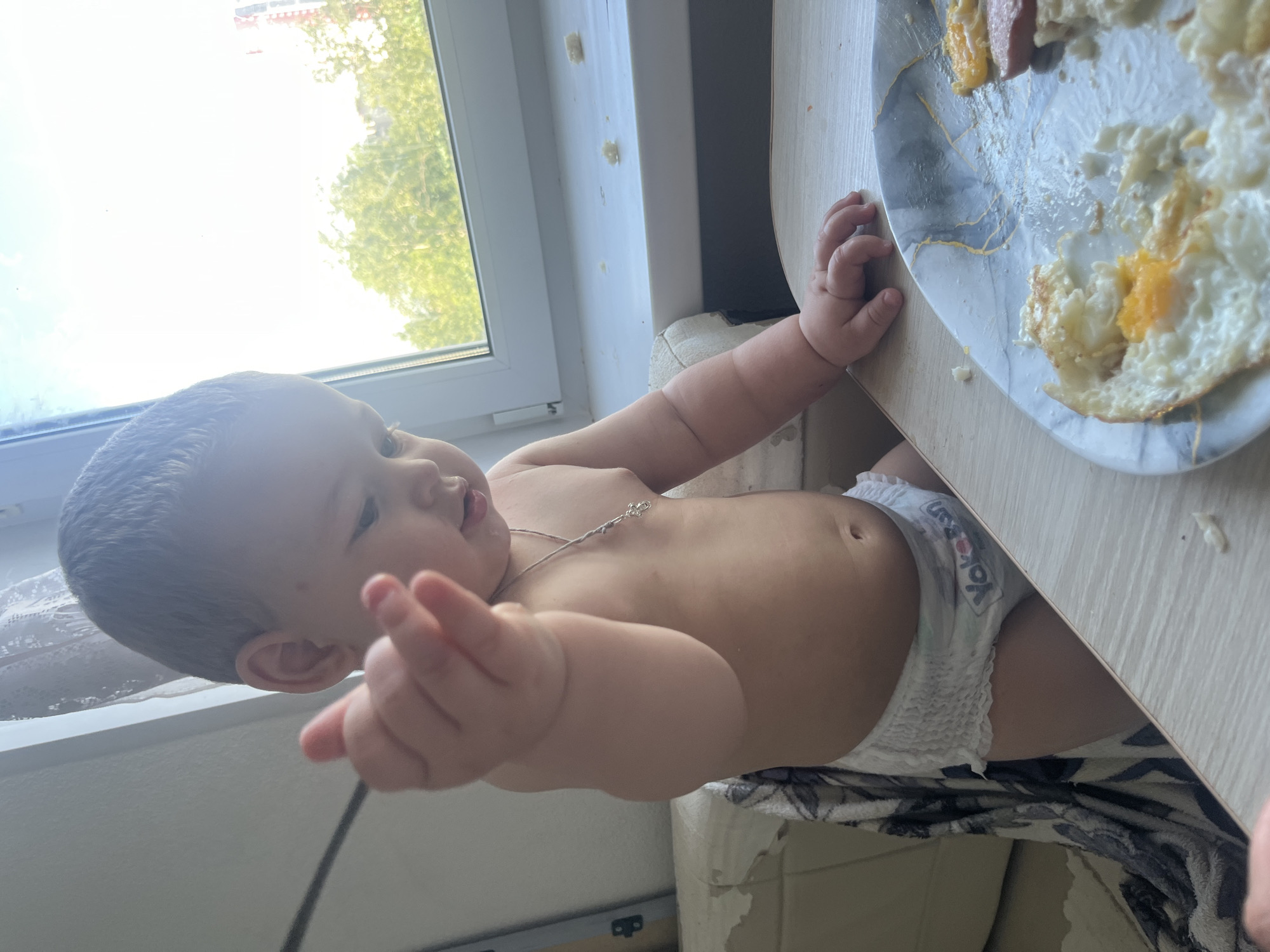 A toddler, wearing a diaper with "YokoSan" branding, is seated at a table. The toddler is looking down at a plate of food which appears to include fried eggs and sausage. A thin chain with a cross pendant is visible around the toddler's neck. The background shows a window with a view of green trees and a building. The scene takes place indoors, likely in a home, in Chelyabinsk, Russia.