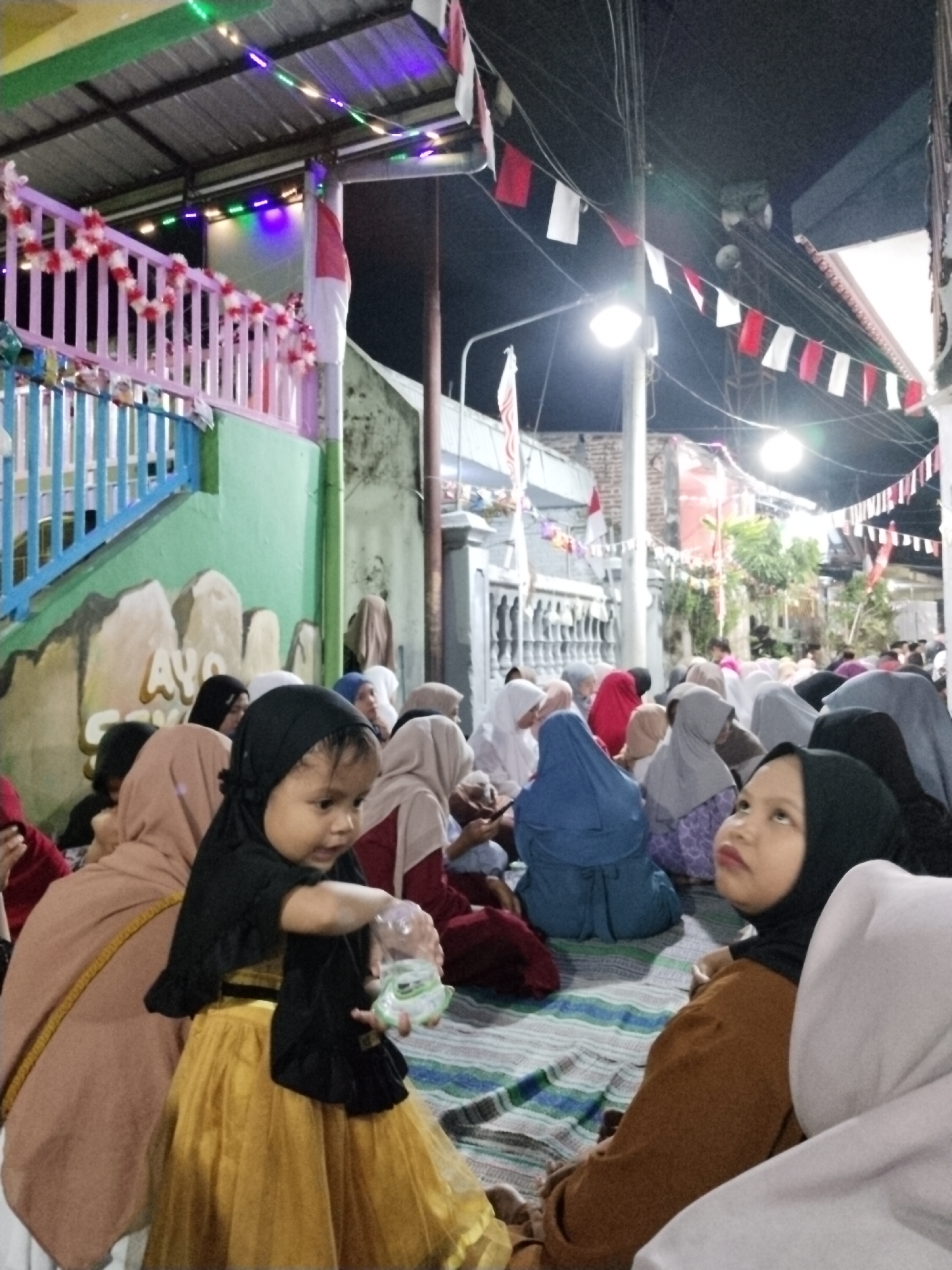 The image depicts an outdoor gathering at night in the city of Ditotrunan, Indonesia. The scene is illuminated by string lights and a bright overhead light. Decorative banners with red and white flags are strung across the area, suggesting a celebratory or nationalistic event. A group of people, mostly women and children, are seated on a striped mat on the ground, facing towards the left of the frame. Many are wearing hijabs and traditional Indonesian clothing. In the foreground, a young child wearing a black hijab and a gold dress is holding a plastic bottle and looking towards the right. The background shows buildings and a decorated railing. The overall atmosphere appears to be one of community and shared experience, possibly during a festival or religious gathering.