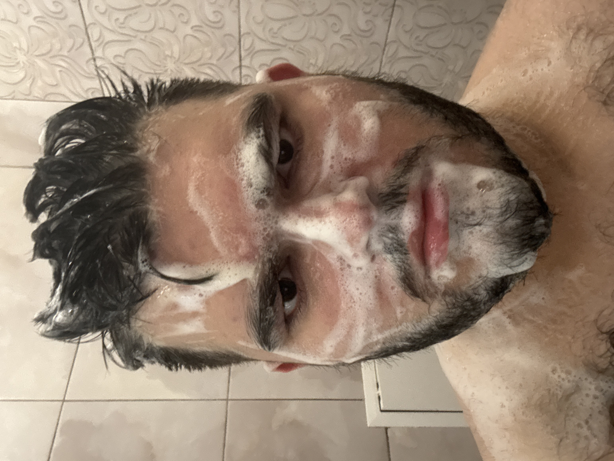 The image shows a person with dark hair and a beard covered in soap or cleanser. The person appears to be in a bathroom, indicated by the tiled walls and shower stall components visible in the background. The focus is on the person's face, with the camera angled upward.
There is no location information in the image; "Burgas, Bulgaria" cannot be confirmed.