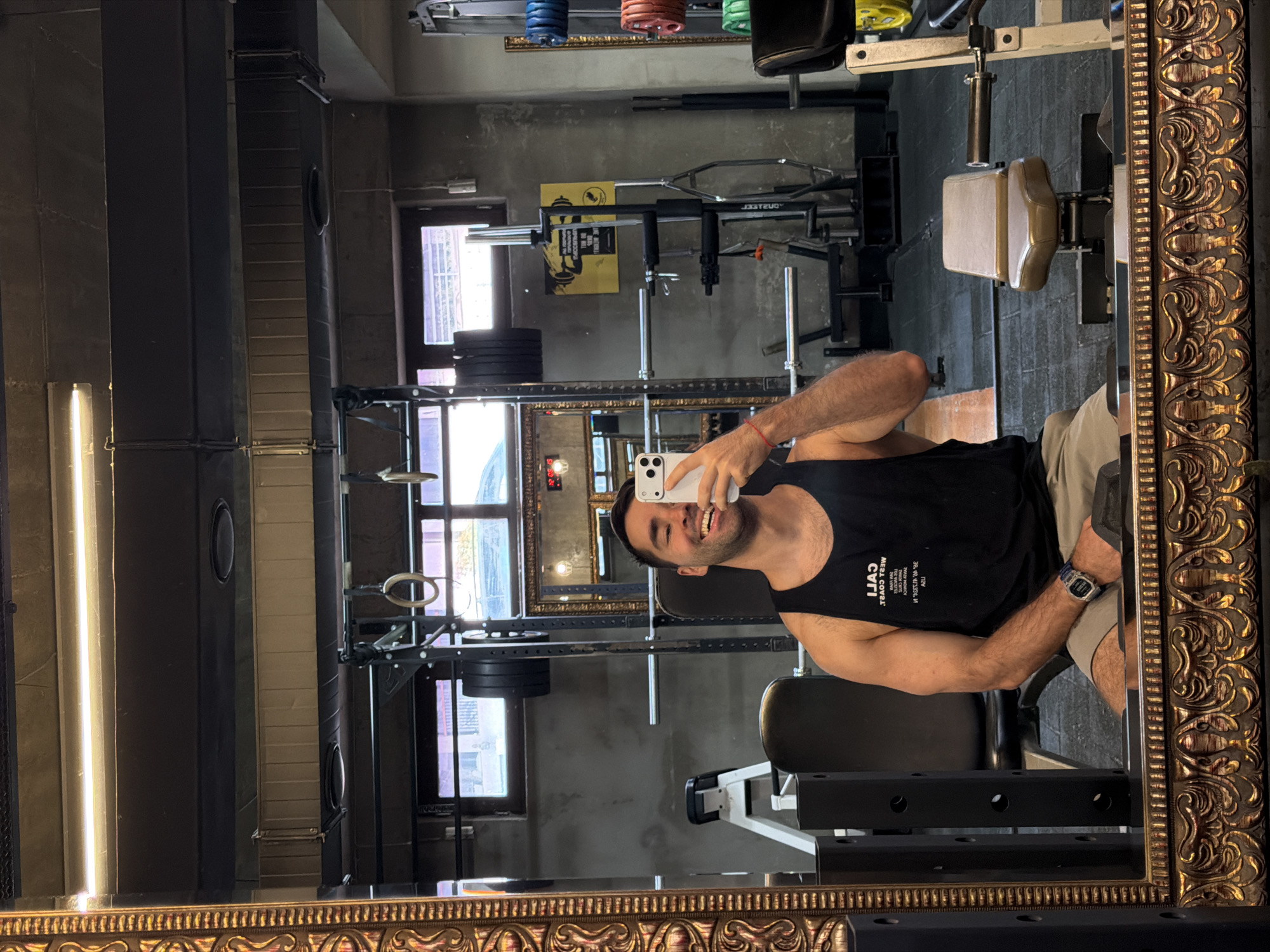 A man takes a selfie in the mirror of a gym in Tbilisi, Georgia. He is fair-skinned with dark hair, wearing a black tank top and khaki shorts, and is smiling. He holds a white smartphone up to his face. The gym is located in an older building with tall ceilings. The gym equipment includes a squat rack, bench press, and weights. The scene appears to be an interior shot, likely taken in the afternoon or evening, with light coming in through the windows.