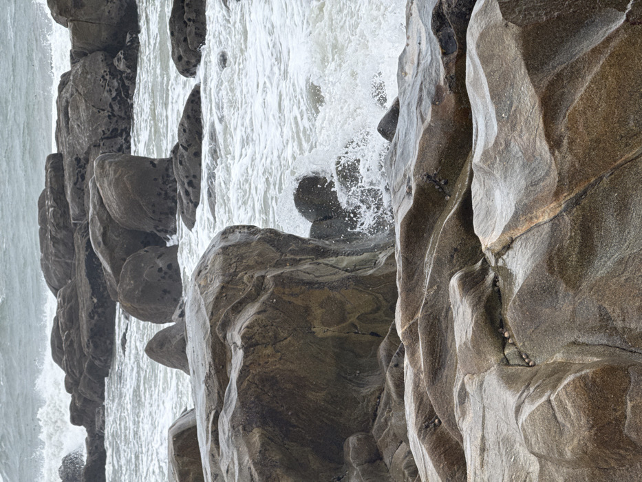 The scene depicts a rugged, rocky coastline in Vila Nova de Gaia, Portugal, under an overcast sky. The foreground and midground are dominated by numerous wet, grey-brown to dark brown rock formations, appearing to be granite or similar metamorphic rock, varying in size and shape. These rocks exhibit smooth, wave-worn surfaces, crevices, and striations. Small clusters of marine organisms, possibly limpets or barnacles, are visible on the exposed rock surfaces in the lower sections of the image.
Ocean waves are actively crashing against and flowing around these rock formations. White sea foam and spray are prominently featured in the midground, indicating significant wave action, particularly around a large, dark rock in the center-left where water is vigorously splashing. The ocean water is a grey-green color, characterized by whitecaps and turbulent conditions extending towards the horizon in the background. No people, animals, or man-made structures are visible. The environment is entirely natural, focusing on the dynamic interaction between the sea and the rocky shore.