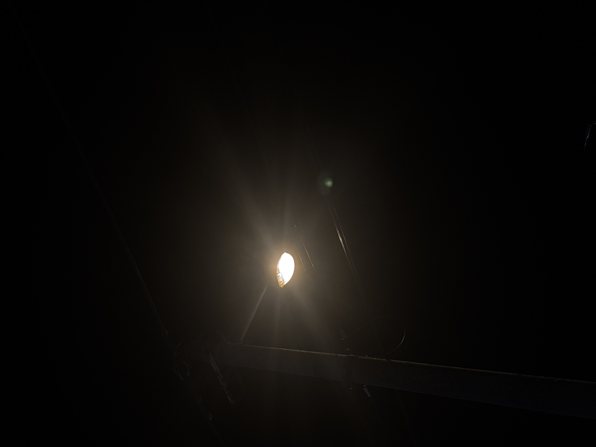 A solitary light illuminates a small segment of a metal structure in an extremely dark environment. The light is centrally located, with beams radiating outward. Parts of the metal structure are barely visible around the light source. The location cannot be determined from this image.