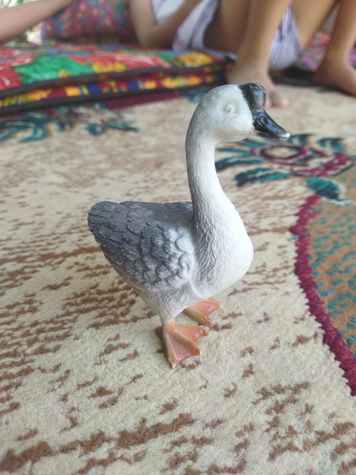 A close-up, eye-level shot captures a toy goose standing on a patterned rug. The goose, primarily white and gray with orange feet and a black and gray head, is positioned slightly to the left of center. Its body is turned to the right, and its head is looking to the right, with one eye visible. The rug beneath the goose features a cream-colored base with brown, abstract markings. In the background, slightly out of focus, are human legs and feet. The legs, one clad in striped fabric and the other bare, suggest the presence of at least two people seated or reclining. Behind the legs, there are colorful cushions or folded fabric with vibrant patterns of red, green, blue, and yellow. The lighting appears natural and diffused. The setting does not provide specific indicators of Mailuu-Suu City, Kyrgyzstan, but the presence of the people and the indoor/outdoor setting with a rug and cushions are discernible.