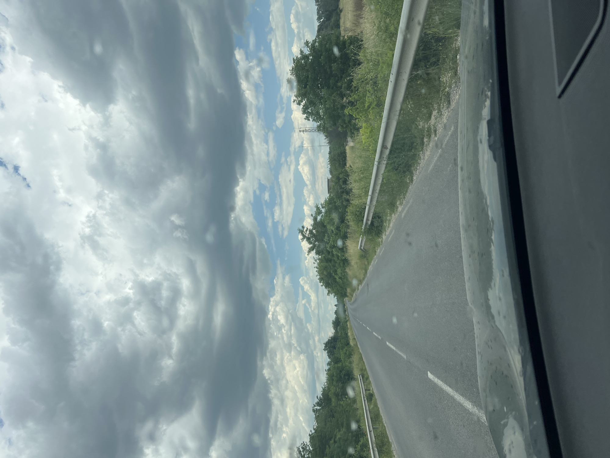 The image shows a view from inside a vehicle, looking out onto a road. The road is asphalt, with white dividing lines, and is bordered by green vegetation and a metal guardrail on one side. The sky is visible with a mix of dark, heavy storm clouds and patches of blue with white clouds. The image appears to be taken while the vehicle is in motion. The location is possibly near Kalekovets, Bulgaria, suggested by the landscape and environmental features.