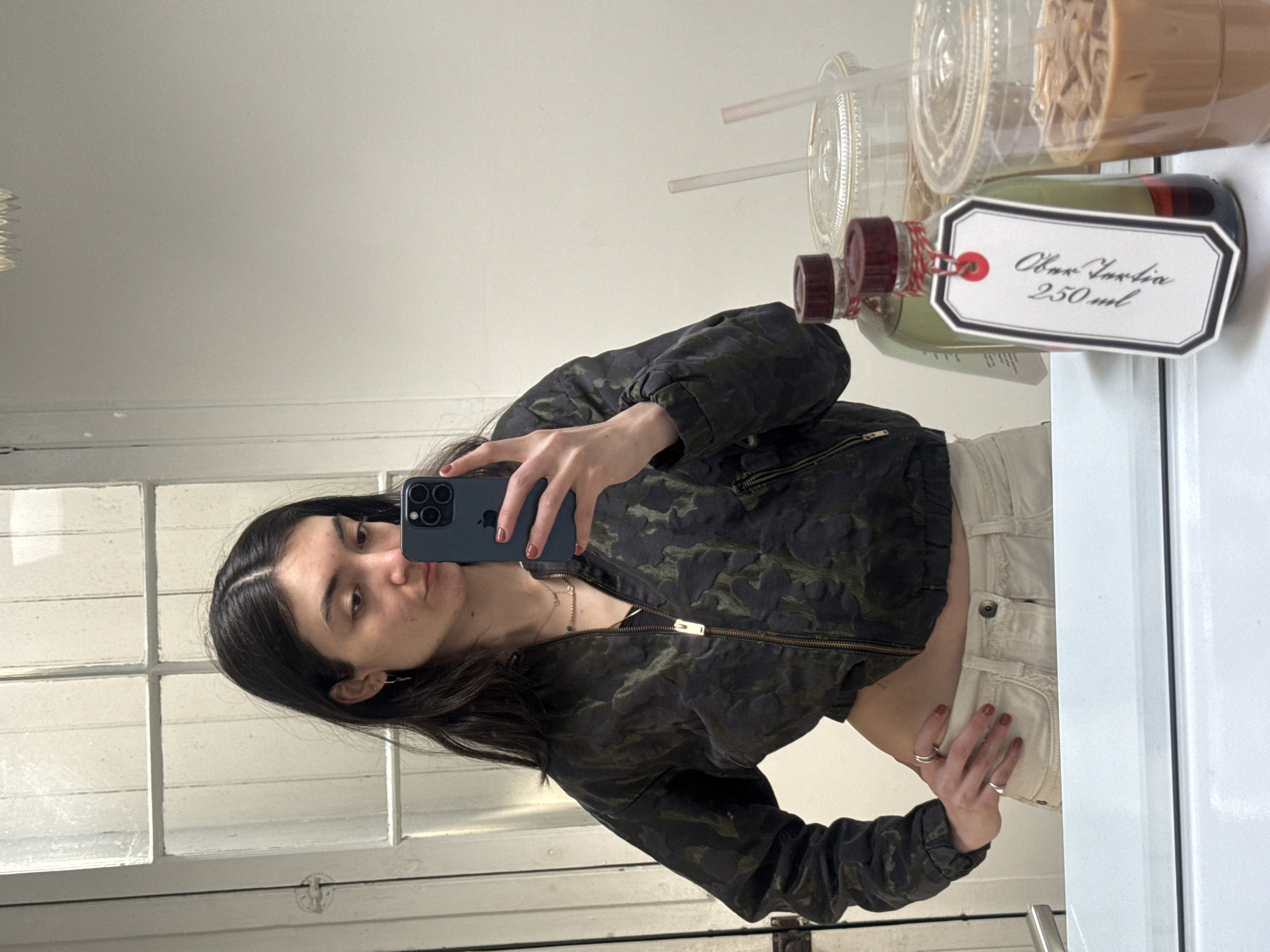 A woman stands in front of a mirror, taking a selfie with a smartphone. She has dark hair and is wearing a patterned jacket and light-colored pants. In the background, there are beverages, including what appears to be an "Oliv Turtia" drink in a 250 ml bottle. The location appears to be indoors, near a window, possibly in New York City, USA, based on the style of the window and architectural elements in the reflection. The woman is the main subject, and her action is taking a self-portrait using her mobile phone.