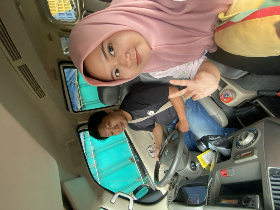 Two people are inside a car. A woman wearing a pink hijab is in the passenger seat, making a peace sign. A man is driving, wearing a black shirt and blue jeans. The car's interior is beige and grey. The car has a sunroof and a view of a structure with blue tarps. A yellow object is visible behind the woman's head.