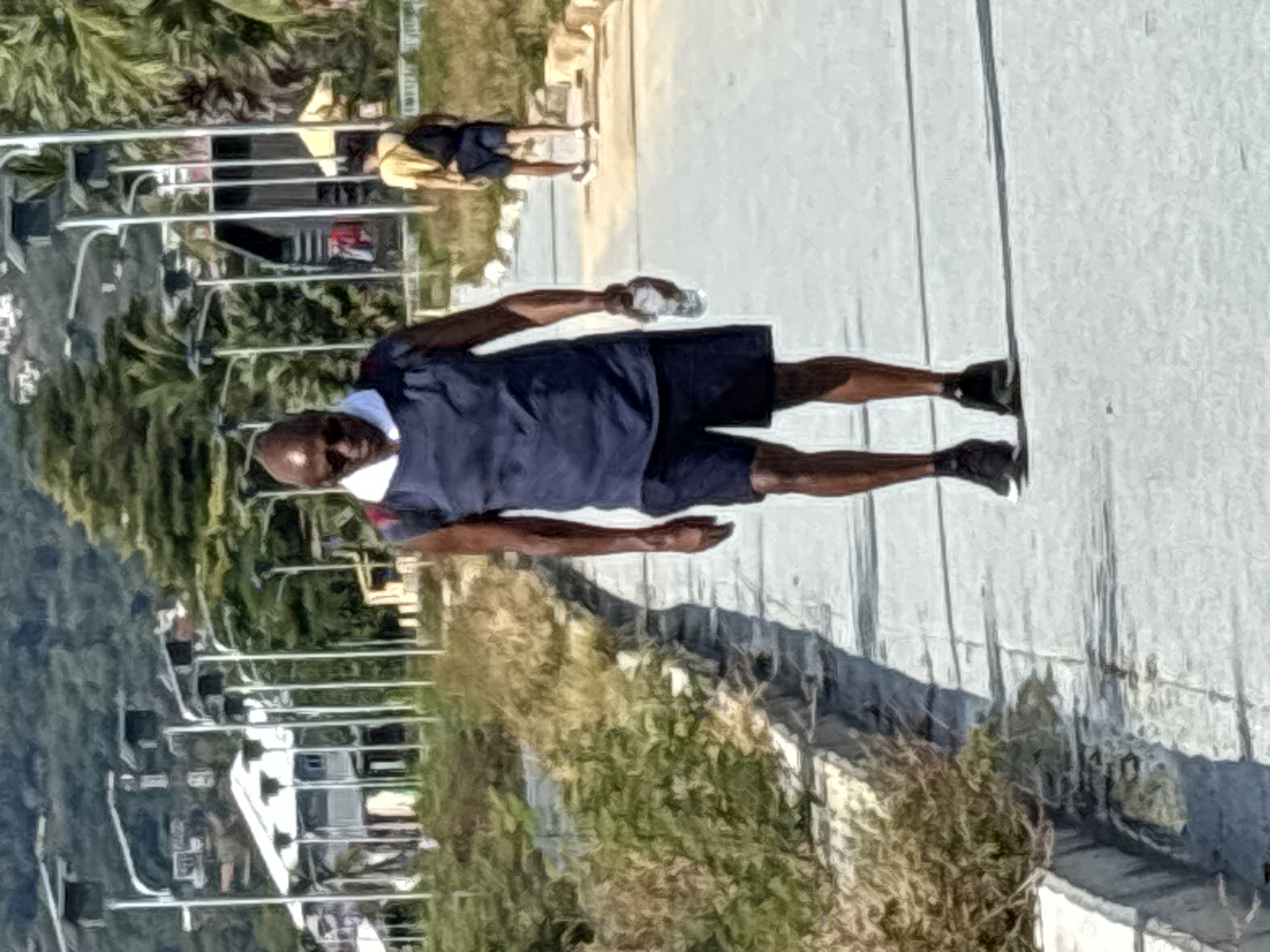 A bald-headed Black male in a blue t-shirt and black shorts walks along a paved pathway near Baan Chaweng, Thailand. He holds a clear water bottle in his left hand. He wears a white face mask. In the background, there are lush green trees and some architectural structures, including possibly a chairlift. Another person is sitting in the background on the paved pathway, beyond the metal bars.