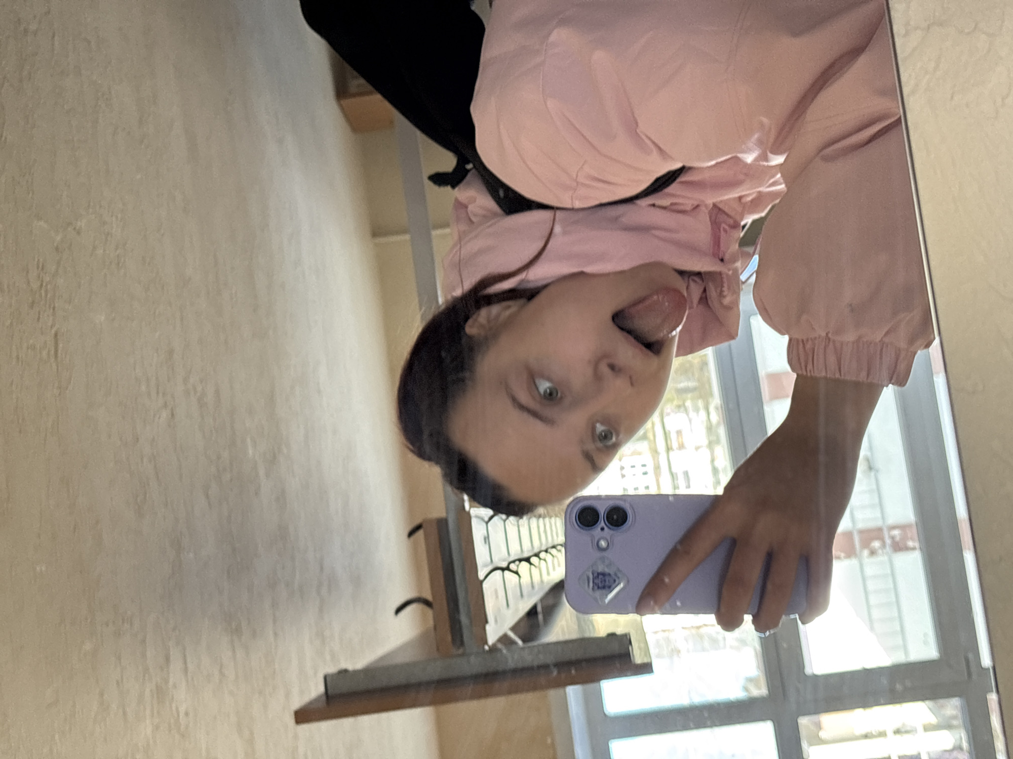 A young woman takes a selfie in a mirror. She is wearing a pink jacket. She sticks out her tongue. She holds a light purple phone in her hand. The reflection in the mirror shows a coat rack and a window. The environment appears to be in a hallway. There is no way to determine the location of the photo in the city of Chaikovsky, Russia.