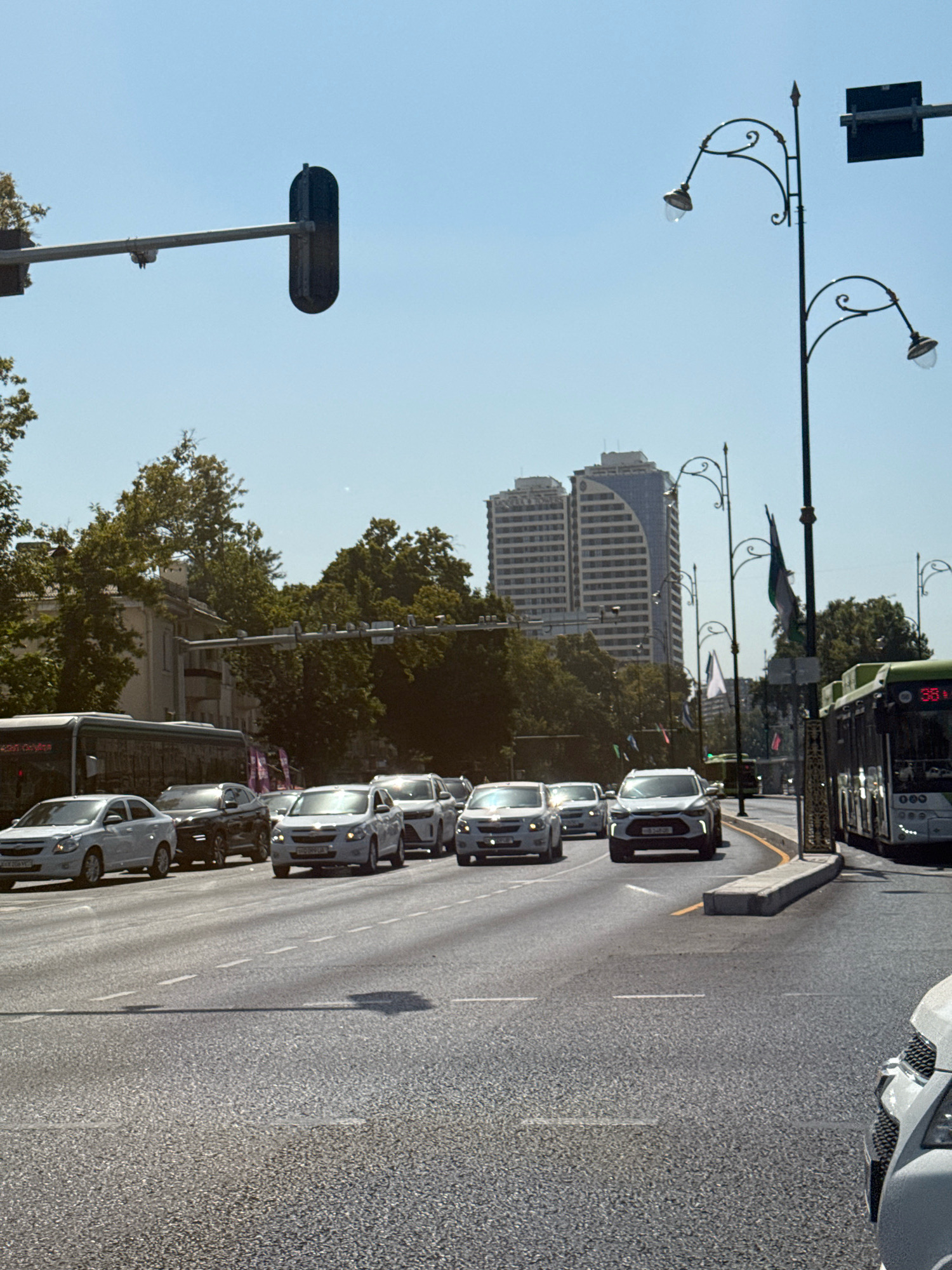 The image depicts a street scene in Tashkent, Uzbekistan, on a clear, sunny day. The perspective is from a vehicle moving along a multi-lane road. In the foreground, the asphalt road surface is visible with dashed white lane markings. Several cars are in motion, including white sedans and a dark SUV. A white SUV is in the center of the frame, with its headlights illuminated. To the right, a green and white bus is traveling in the same direction. In the background, a row of trees lines the street, with modern, multi-story buildings visible behind them, including two distinctive twin towers with a curved facade. Ornate street lamps with decorative arms and glass shades are present along the roadside, some with flags attached. Traffic signals are visible overhead, including a prominent black traffic signal housing on a metal arm. The sky is a pale blue, indicating good weather. The scene suggests a busy urban environment.