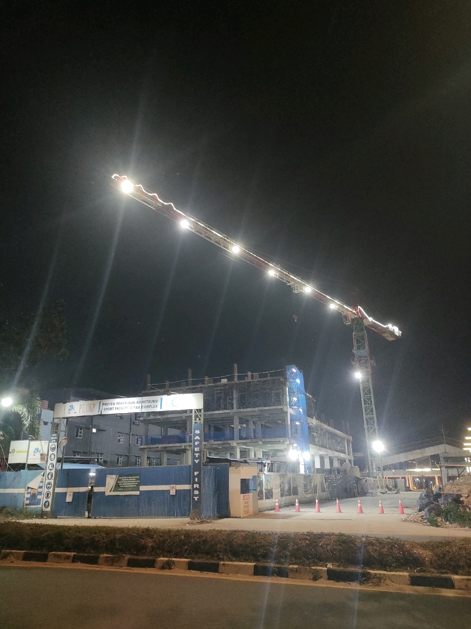 The image depicts a construction site at night, likely in Batam, Indonesia, as indicated by the signage. A large tower crane dominates the scene, its boom illuminated by numerous lights. The crane is positioned over a multi-story building under construction, which appears to be a sports facility and F&B complex based on the Indonesian text on the signage.
The building itself is a concrete structure with exposed rebar and scaffolding, indicating ongoing work. Various construction materials are visible, including piles of rubble and construction fencing. Orange traffic cones are placed in a line across the site.
A prominent sign at the entrance reads "PT. OKP PROYEK PEKERJAAN KONSTRUKSI SPORT FACILITY & F&B COMPLEX". Another sign on a vertical structure states "SAFETY FIRST" in large, vertical letters.
The foreground shows a road with a concrete median and grass. The overall impression is one of a bustling construction project operating through the night, with artificial lighting illuminating the work area against the dark sky. No people are clearly discernible in the image.