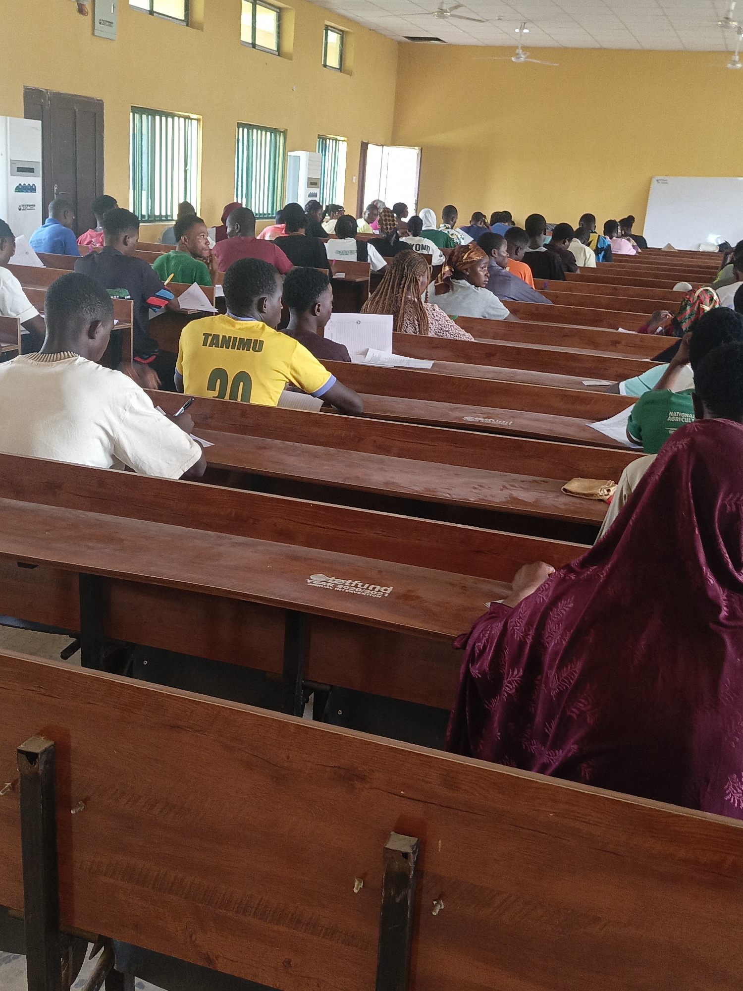 In Akwanga, Nigeria, a medium shot shows a classroom filled with students seated at rows of wooden desks with black metal frames. Most students face forward; a few look sideways or at papers on their desks. A man in a yellow shirt with "TANIMU" and "00" printed on the back is prominent. Some students are writing, and papers are visible on desks. The classroom has yellow walls, barred windows, and ceiling fans. A whiteboard is on the right, and a utility box is on the left.