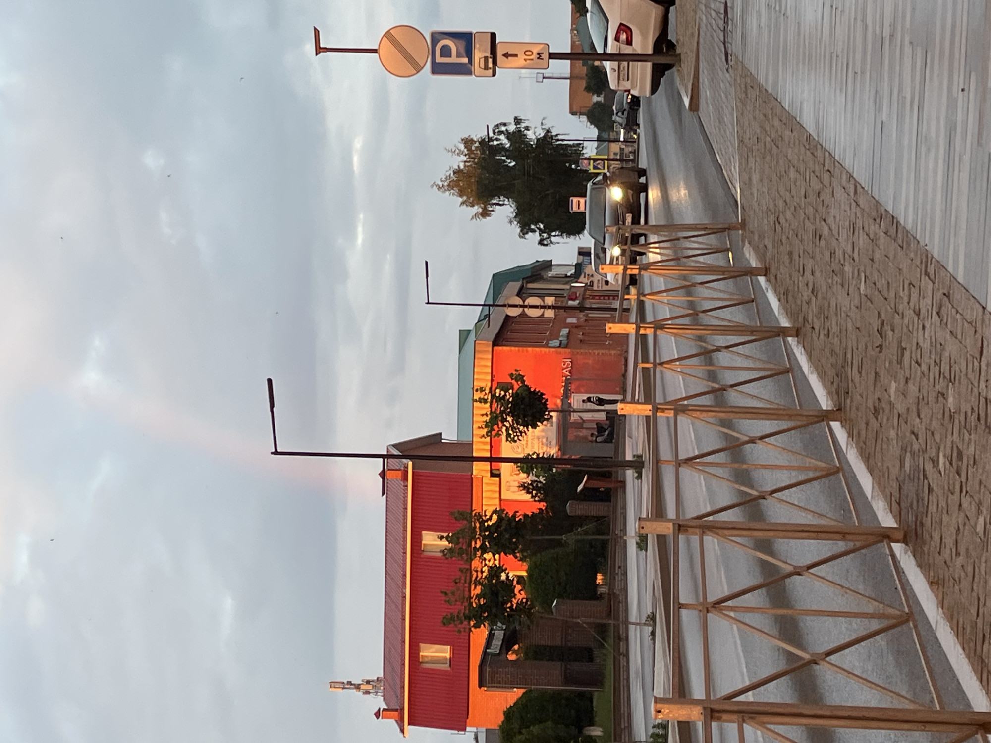 The image depicts a street scene in Kukmor, Russia, under an overcast sky with a faint rainbow visible. The foreground features a sidewalk paved with interlocking bricks, bordered by a decorative metal fence with a diamond pattern. A paved road runs alongside the sidewalk.
On the left side of the road, a building with a bright red, pitched roof and orange lower walls is illuminated by the setting sun. Several trees are situated in front of this building. In the background, a communication tower is visible.
On the right side of the road, a traffic signpost displays several signs: a circular sign with a diagonal red bar indicating the end of a restriction, a blue square sign with a white 'P' denoting parking, and a rectangular sign with a car symbol, an upward arrow, and "10 M" indicating a parking distance. Further down the road, vehicles are present, including a white car parked on the right and several cars driving on the road, some with their headlights on. A large tree with dense foliage stands near the traffic signpost. The overall impression is that of a street in a town during the late afternoon or early evening.
