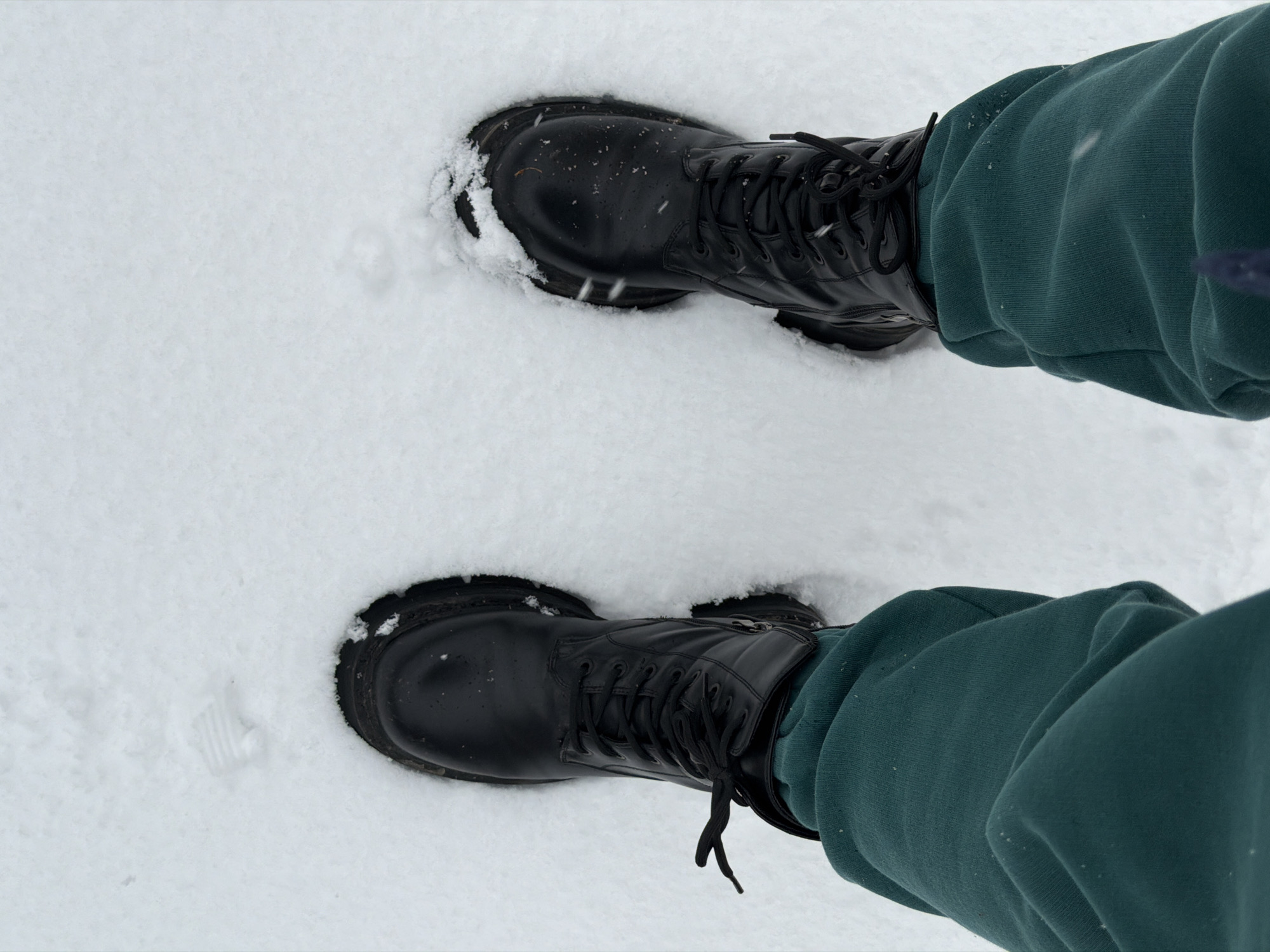 The image shows a view from above of two black leather boots standing in the snow. The boots are laced and have thick soles. The person wearing the boots is also wearing teal-colored sweatpants. The environment is a snowy area. The location is in Chaikovsky, Russia.