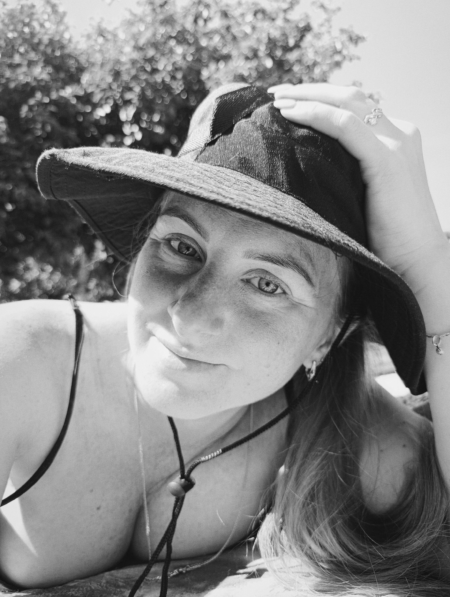 A close-up, black and white photograph features a young adult female in an outdoor setting. The subject is oriented towards the camera with her head slightly tilted, her gaze directed forward, and a faint smile present on her face. Freckles are visible across her nose and cheeks. She wears a dark, wide-brimmed hat, with her right hand gently resting on the brim near her temple. A decorative ring is visible on her right ring finger, and a delicate bracelet with a small charm adorns her right wrist. Her long, light-colored hair extends downwards on the right side of the frame. She wears a dark spaghetti strap top or bikini top, and a dark cord necklace with a visible knot is around her neck/chest. The background is heavily blurred, showing indistinct bright and dark circular patterns consistent with sunlight filtering through foliage, indicative of an outdoor, sunny environment. The image exhibits a subtle grain. Location: Unknown, Unknown.