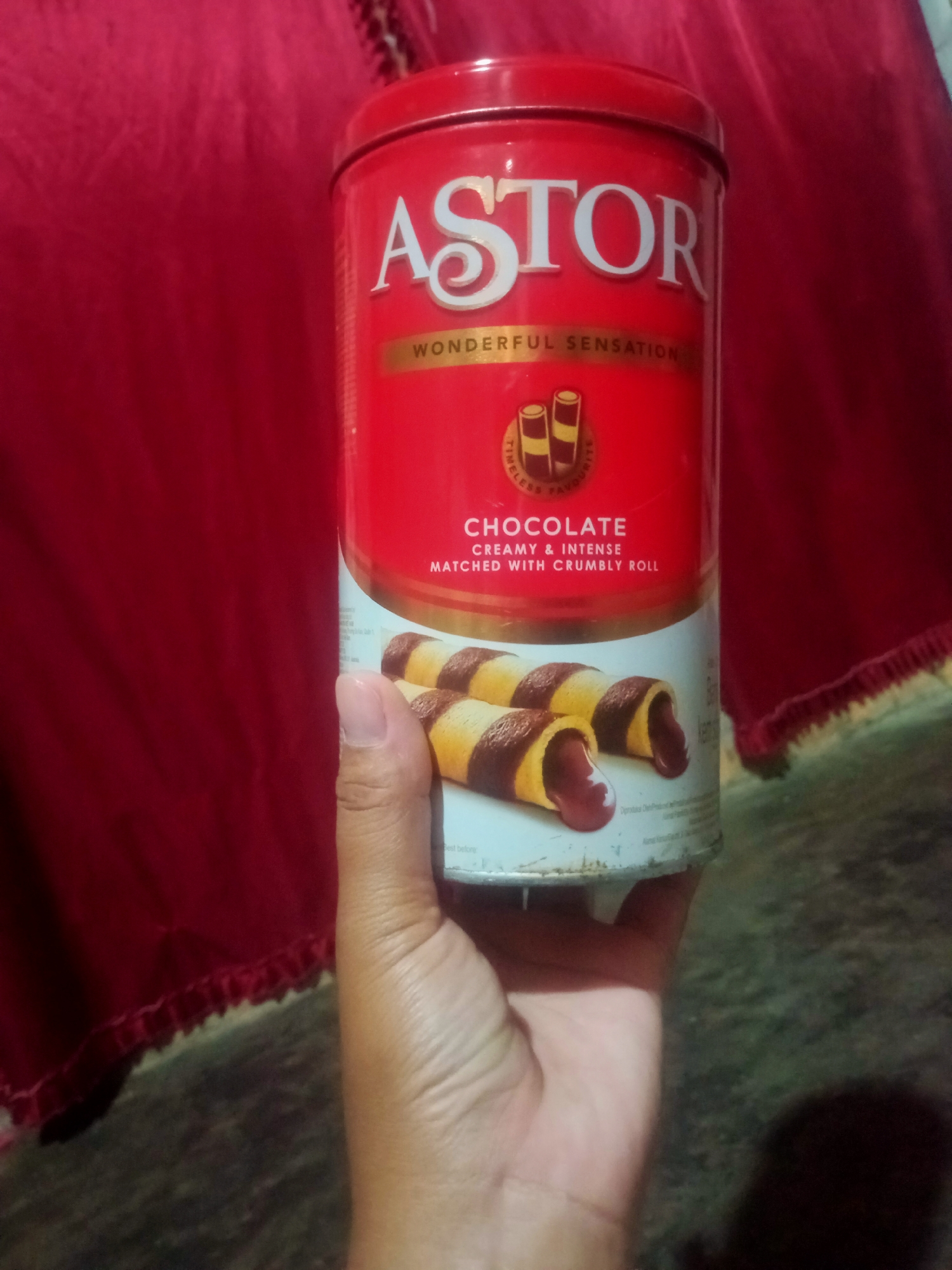 A hand holds a red cylindrical tin of Astor chocolate wafer rolls. The tin has white text that reads "ASTOR" at the top, with smaller text below it saying "WONDERFUL SENSATION". There is an image of two chocolate wafer rolls on the front of the tin, showing chocolate filling oozing out. The background is a dark red fabric, possibly a tablecloth or drapery. The image appears to have been taken indoors, but there is no specific information to place it in Nusa Agung, Indonesia.