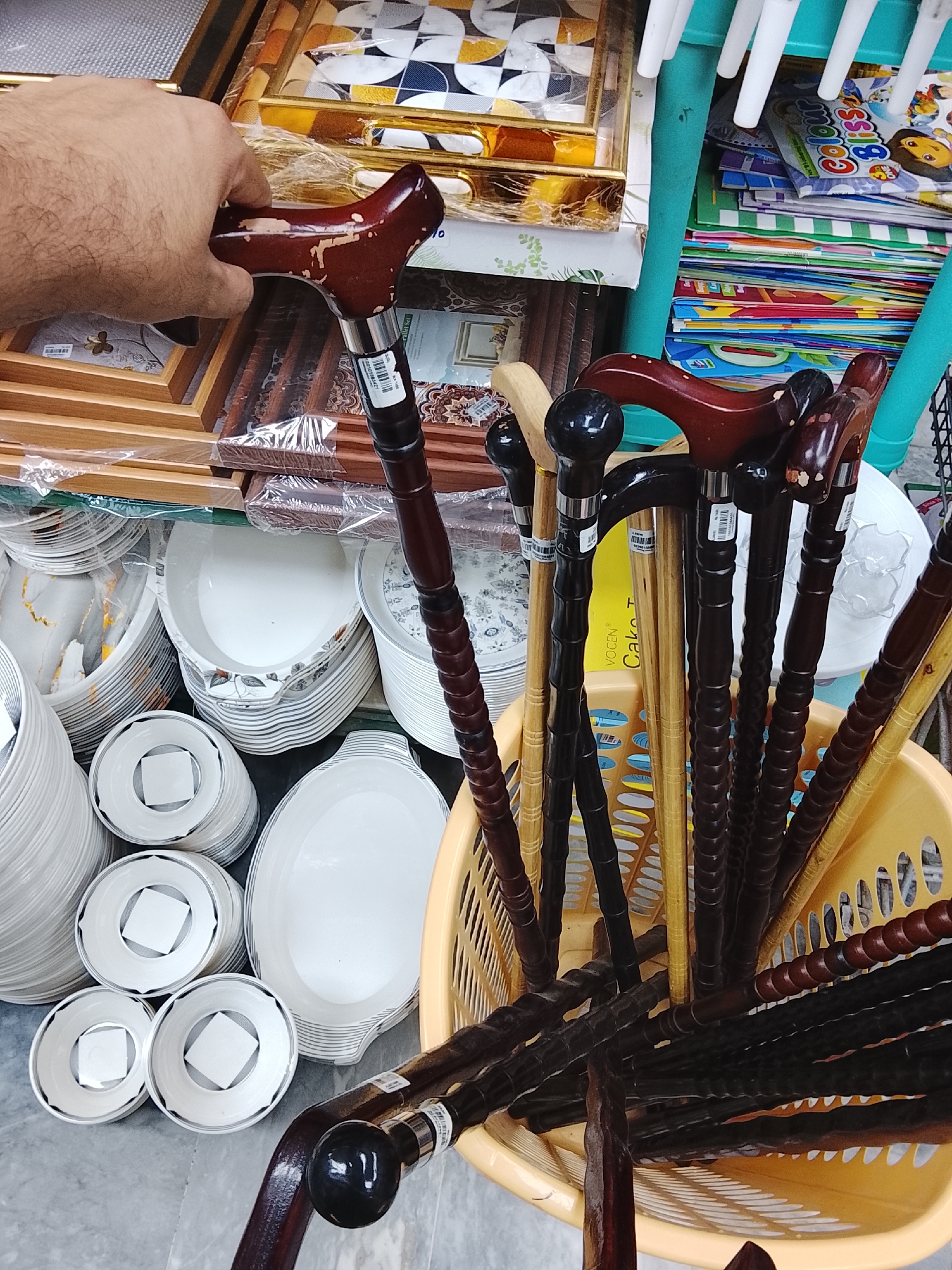 A photograph taken in Gulgasht Colony, Pakistan, shows a diverse collection of items, including walking canes, dishware, and picture frames. A hand is shown holding one of the walking canes. The canes, of varying colors and styles, are stored in a yellow plastic basket. To the left of the basket, stacks of white dishes are arranged, including plates, bowls, and serving dishes. On a shelf above, a selection of picture frames are displayed. Additional objects such as coloring books are shown in the background. The image appears to have been taken indoors, in a retail setting.