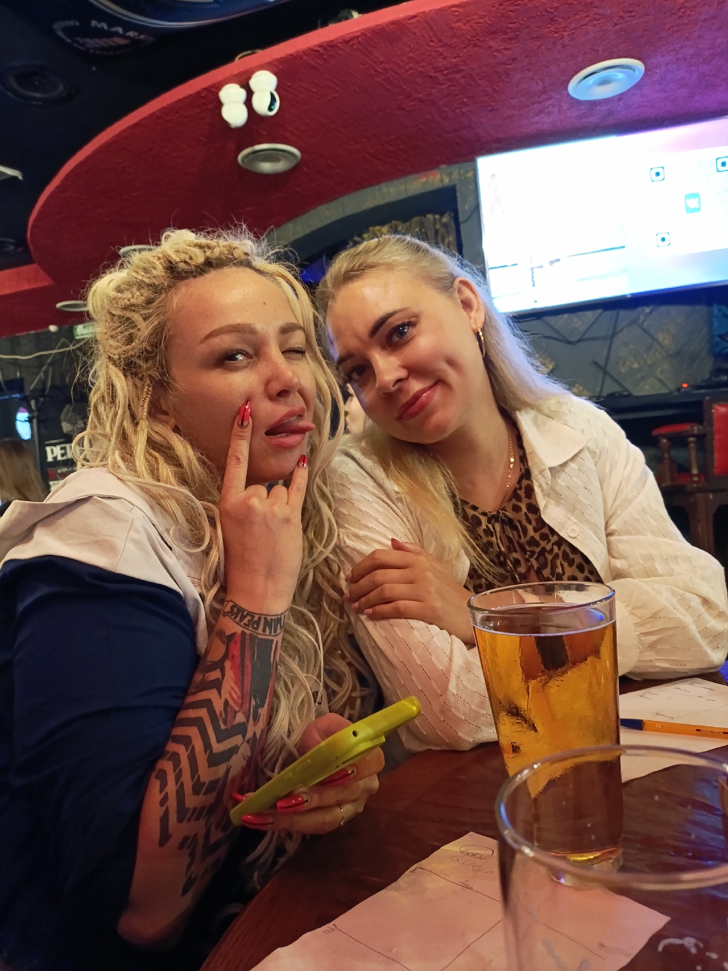 Two women are sitting at a table in a dimly lit establishment, possibly a bar or restaurant. The woman on the left has blonde, curly hair and is wearing a blue and gray hooded jacket. She is sticking out her tongue and making a "rock on" hand gesture, with long red nails. She has a large tattoo on her arm with black, geometric patterns and some red coloring. She is holding a bright yellow object, possibly a phone case. The woman on the right has blonde hair pulled back and is wearing a white button-up shirt with a leopard print top underneath and a gold necklace. She is smiling gently at the camera. Both women appear to be in good spirits.
On the table in front of them are two glasses of beer, one of which is full and golden. There are also some papers and pens on the table. In the background, a large screen is visible, displaying a game interface with icons and graphics. The ceiling above them is red and curved, with some lights and decorative elements. The overall atmosphere suggests a casual outing, perhaps during the evening. The location is identified as Chaikovsky, Russia.