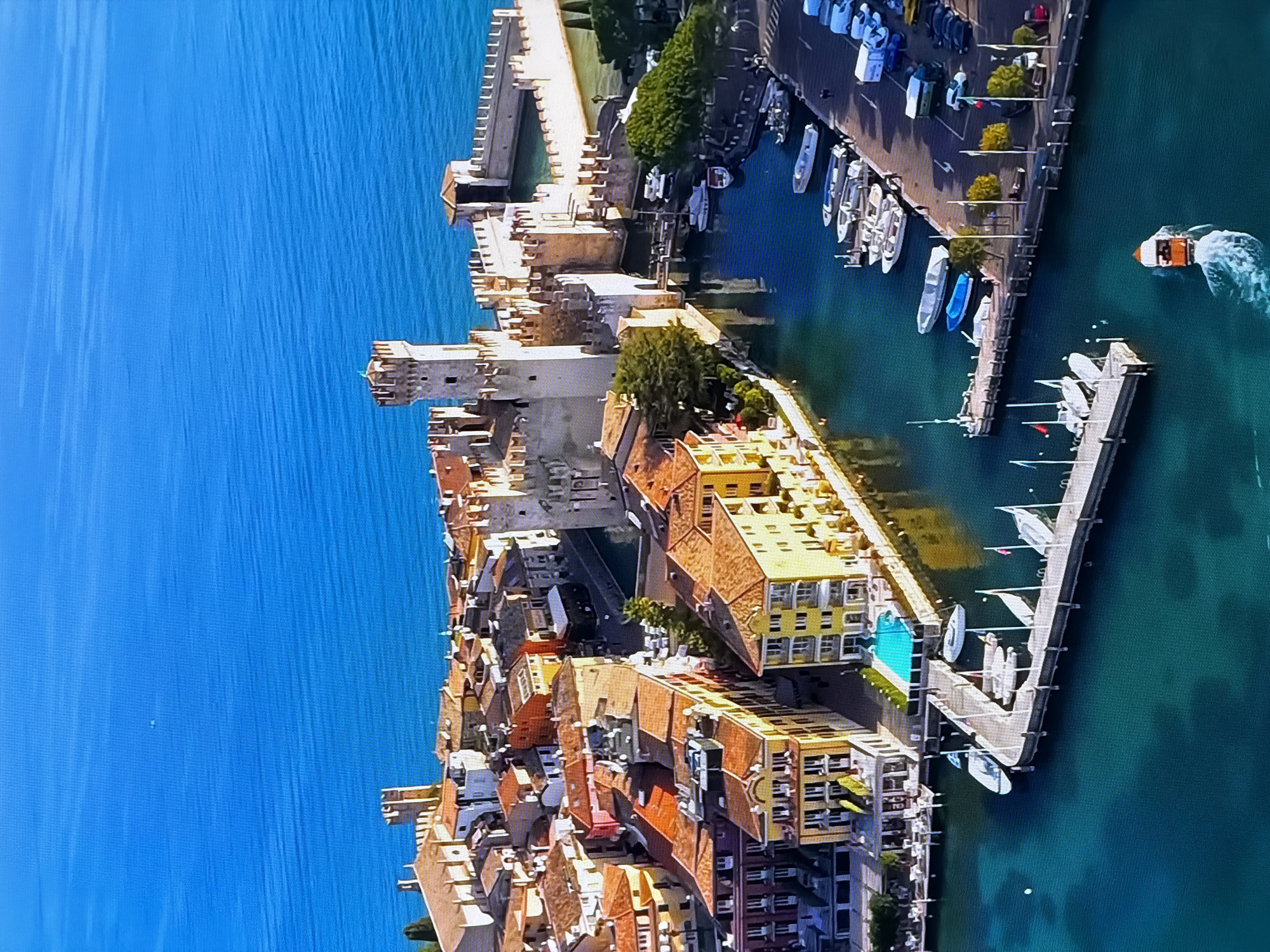 This is an aerial, high-angle shot of a picturesque harbor town with a fortified castle and a body of water. The water is a vibrant blue, with ripples suggesting light wind. Several boats, ranging from small speedboats to larger yachts, are docked along the piers and in the harbor. The town features buildings with terracotta roofs and colorful facades, lining canals and streets. A prominent stone castle, with crenellated towers and thick walls, stands at the water's edge, seemingly integrated with the town. The scene is bathed in bright sunlight, casting shadows and highlighting the architectural details. There are a few people visible on the docks and near the buildings. The overall impression is one of a charming, historic European lakeside settlement. The location is not Chirchik, Uzbekistan.