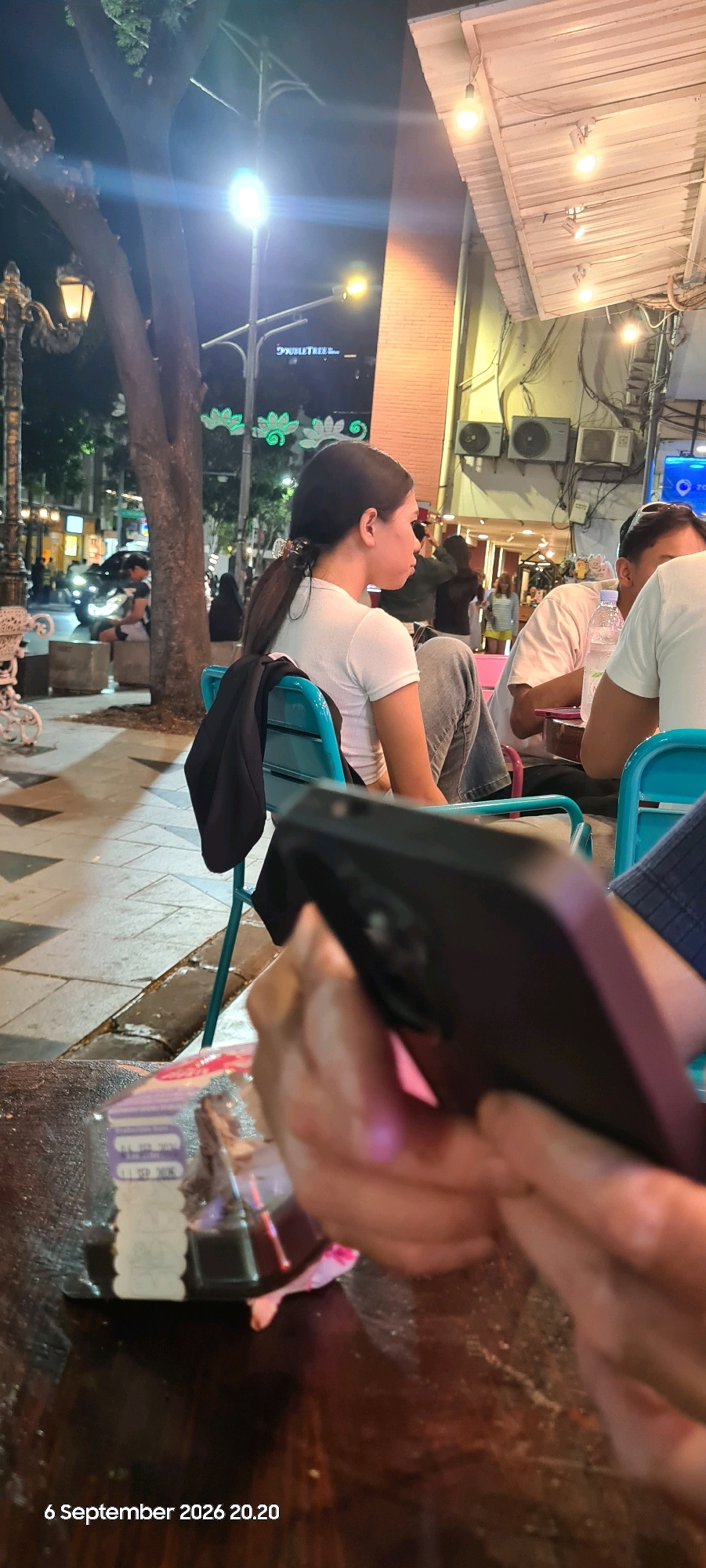 A side-view of a young woman with dark hair pulled into a ponytail is captured from behind, seated at an outdoor table at night. She is wearing a white t-shirt and jeans, with a dark jacket draped over the back of her teal chair. In the foreground, a person's hands hold a dark-colored smartphone, partially obscuring a wooden table where a small plastic container with food and some labels are visible. The background reveals a dimly lit street scene in Surabaya, Indonesia, with trees, streetlights, and brick buildings. A sign for "DOUBLETREE" is faintly visible on one of the buildings, and traffic can be seen in the distance. The timestamp "6 September 2026 20.20" is overlaid on the bottom left of the image.