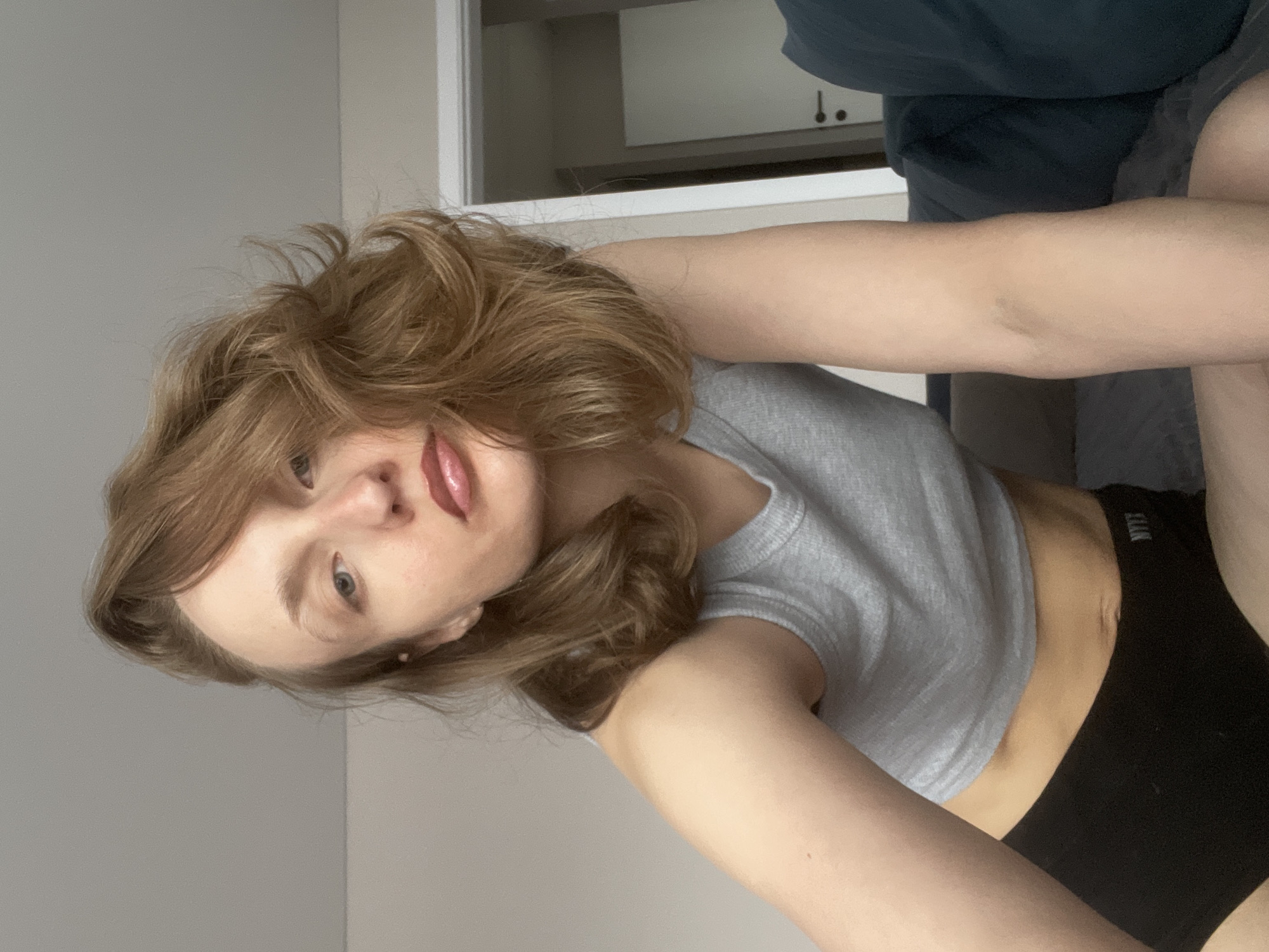 A young woman with light brown hair is facing the camera. She is wearing a gray crop top and black underwear. She appears to be indoors, possibly in a bedroom. In the background, there is a doorway or closet. The location of the image is not identifiable as Подшивалово, Russia.