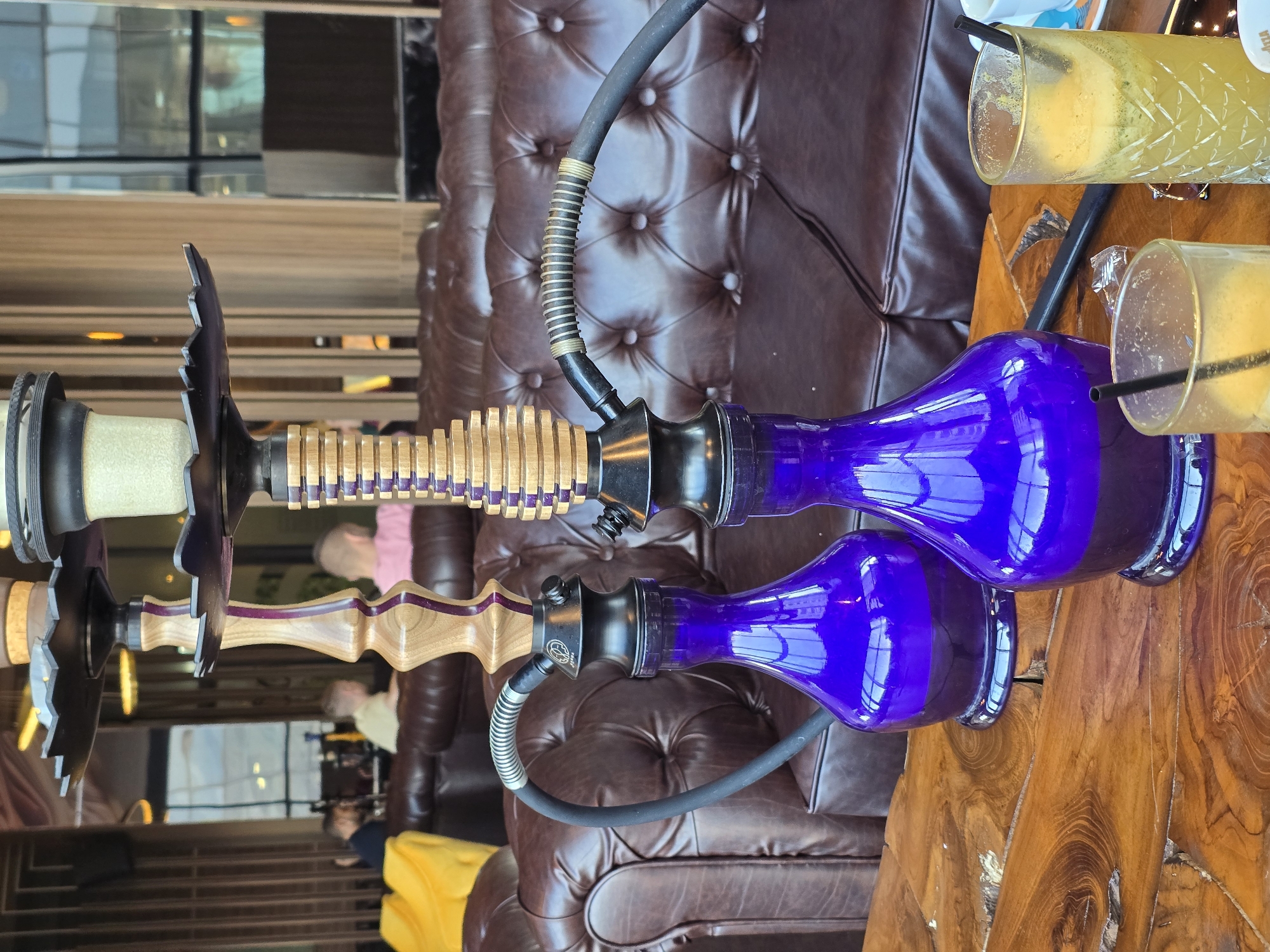 Two hookahs are on a wooden table in what appears to be a lounge or bar. The bases of the hookahs are a vibrant purple. Each hookah has a hose attached. Two glasses with straws are partially visible on the table next to the hookahs. The background shows a tufted leather couch and a blurred interior with people and other objects, suggesting the lounge is in use. The location is Burgas, Bulgaria.
