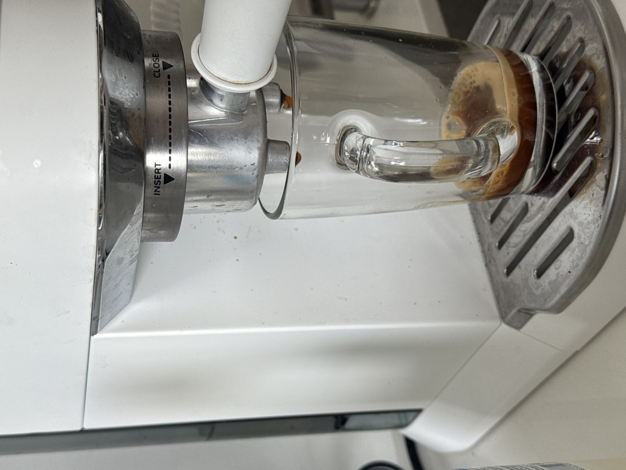 The image shows a close-up of an espresso machine dispensing coffee into a glass cup. The machine is primarily white and silver, with markings indicating "INSERT" and "CLOSE" on a rotating mechanism. A clear glass cup with a handle is positioned to catch the dispensed coffee, which is visible as a dark liquid with some froth. The background is a plain white surface. The location is not discernible from the image.