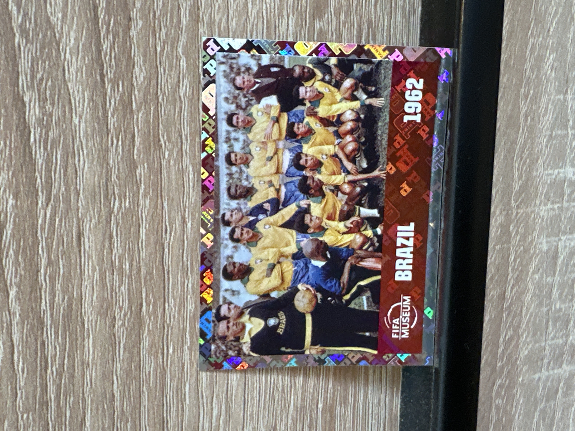 The image shows a rectangular trading card featuring a photograph of the Brazil national football team from 1962. The card is placed on a wooden surface with a visible grain pattern. The photograph depicts a group of men in football kits, some standing and some kneeling, posing for a picture. The card has holographic borders. Along the bottom edge of the card, there is text that reads "FIFA MUSEUM BRAZIL 1962". There are no discernible elements in the image that would suggest a location in Prisovo, Bulgaria, or any specific city. The scene captured in the photograph is a sports team photo taken outdoors, likely on a football field.