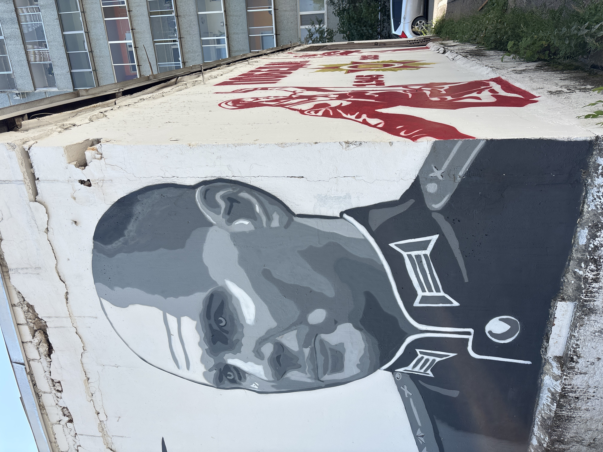 A mural of a man in a dark military uniform is painted on the exterior wall of a building. The man's face is rendered in shades of gray, with his head shaved and his gaze directed to the left. The uniform features epaulets with white detailing and a circular emblem on the chest. To the right of the portrait, on an adjacent section of the wall, is a red stencil of a figure, possibly a soldier, alongside text and a golden star. This mural is located on the side of a building in Blagoveshchensk, Russia, adjacent to a grey, multi-story apartment building and a patch of overgrown grass. A silver car is parked in the background. The wall itself appears weathered, with cracks and some damage visible.