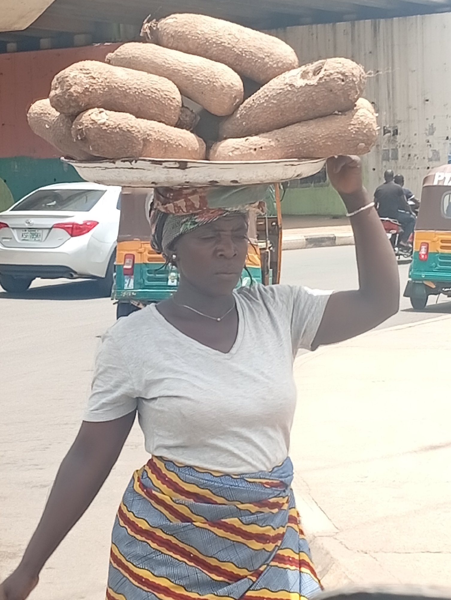 A woman balances a large, round, silver-colored tray piled high with brown, root-like vegetables on her head. She wears a patterned headscarf, a gray v-neck t-shirt, a long wraparound skirt with blue, yellow, and red stripes, and a silver necklace. Her right arm is raised, with a white bracelet on her wrist. She is standing in an urban street setting, as evidenced by the white car, green and yellow auto rickshaws, and two people on a motorcycle in the background. The woman is likely a street vendor. The scene appears to be in Jos, Nigeria based on the auto rickshaw, which is a common form of transport there.