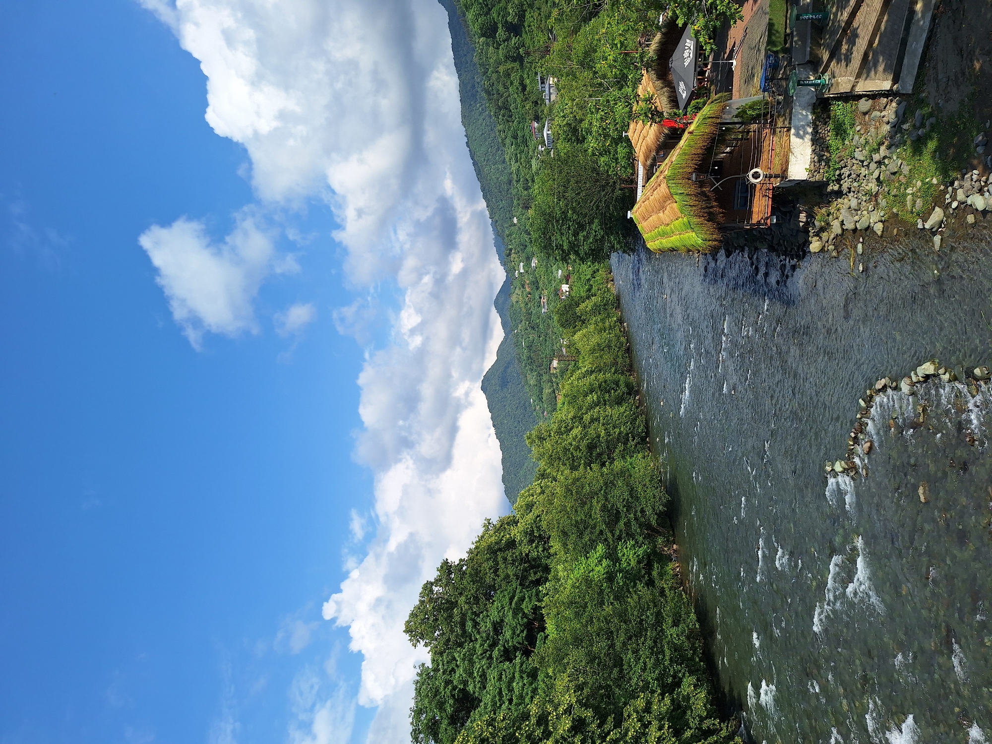 A scenic view of the Chorokhi River in Kobuleti, Georgia, with lush green hills in the background and a clear blue sky dotted with white clouds. On the right bank of the river, there are several rustic-style restaurants with thatched roofs, situated along a rocky embankment. One of the restaurants has a sign with a white lifebuoy hanging on it. The river flows from left to right, with visible currents and ripples. The surrounding vegetation is dense and green, characteristic of a subtropical climate. The image appears to have been taken from an elevated position, possibly a bridge or a higher vantage point overlooking the river and the establishments.