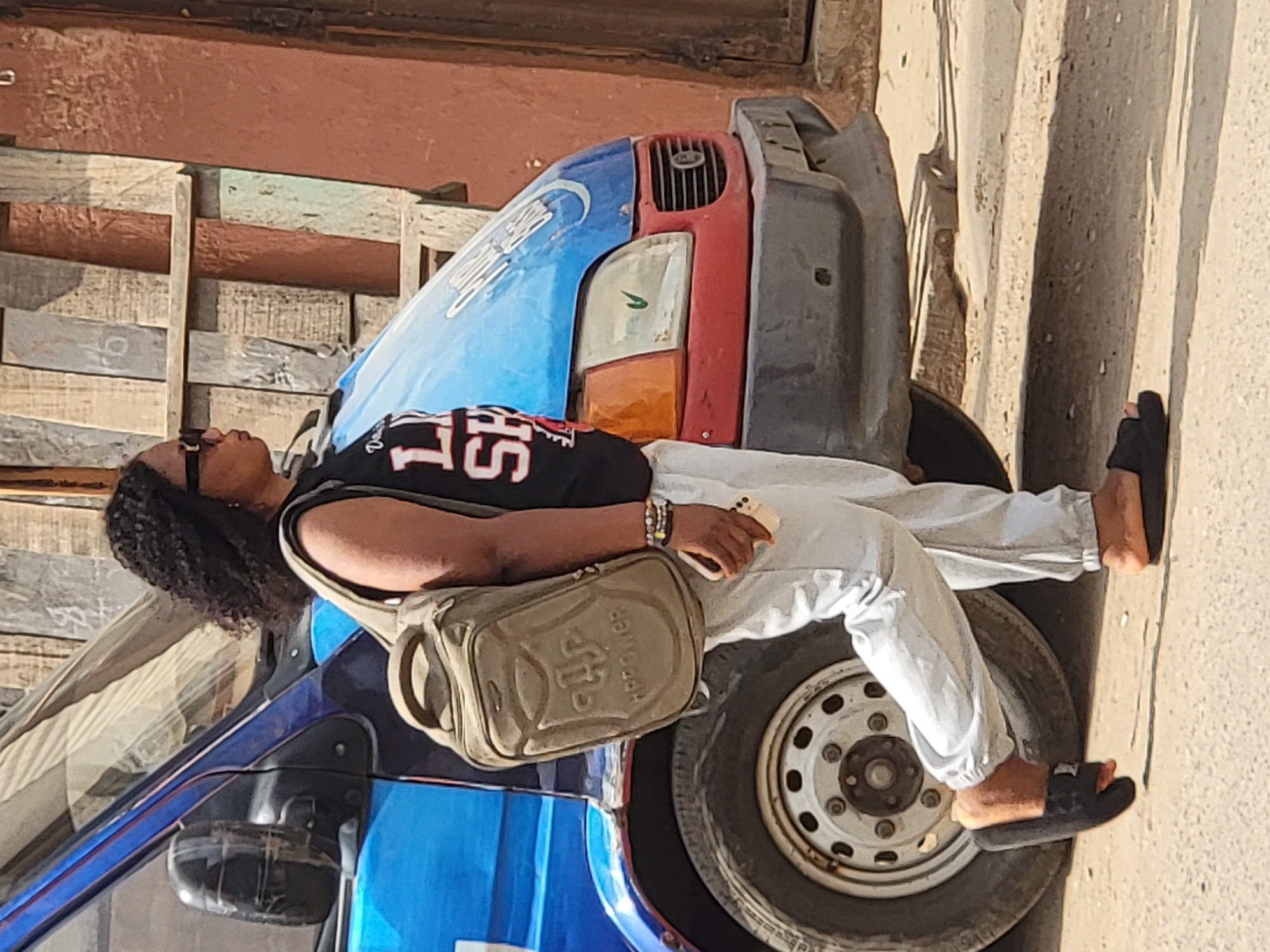 A person is depicted in bright daylight, leaning against a three-wheeled vehicle in what appears to be an urban or semi-urban setting in Maiburiji, Nigeria. The individual, likely a woman with dark curly hair, is positioned on the side of the blue and red patched vehicle, with her right arm resting on its roof and her legs extended to the side. She wears black sunglasses, a black sleeveless top with white lettering "LS" and possibly "15" or similar numbers, light-colored long pants, and black slides/sandals. A tan-colored shoulder bag, featuring an emblem and possibly the text "KOROPTER", hangs across her body. In her left hand, she holds a light-colored smartphone.
The three-wheeled vehicle has a distinctive, customized front featuring blue, red, and dark grey sections, with a visible headlight and grille. The wheel and tire are prominent in the lower right of the image. The background consists of a weathered wooden or plank-built structure, suggesting a building or wall, with varying shades of brown, grey, and faded colors. The ground is a light, dusty-looking surface, indicative of a street or roadside. The strong sunlight and shadows suggest it is midday or early afternoon. The overall scene conveys a casual moment of rest or waiting.