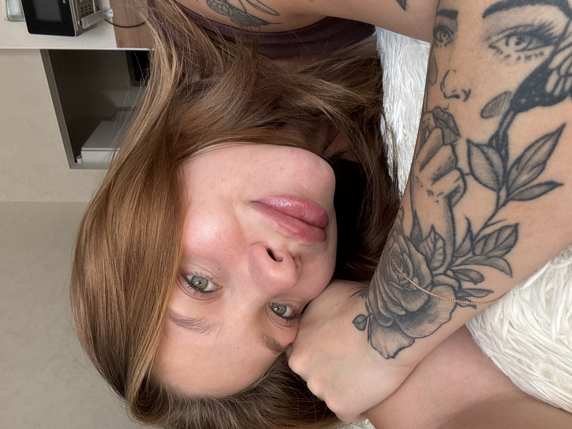 A young woman with auburn hair and green eyes is lying on a fluffy white surface, possibly a rug or blanket, with her head resting on her arm. She is looking directly at the camera with a relaxed and intimate expression. Her left arm, adorned with a large rose tattoo and other designs, is visible, supporting her head. The background is softly blurred, revealing a wall and a shelf with some objects, including what appears to be a small microwave or electronic device. The lighting suggests an indoor setting, possibly a bedroom. There are no visible landmarks or elements in the image that would allow for a specific location within Krasnodar, Russia to be identified.