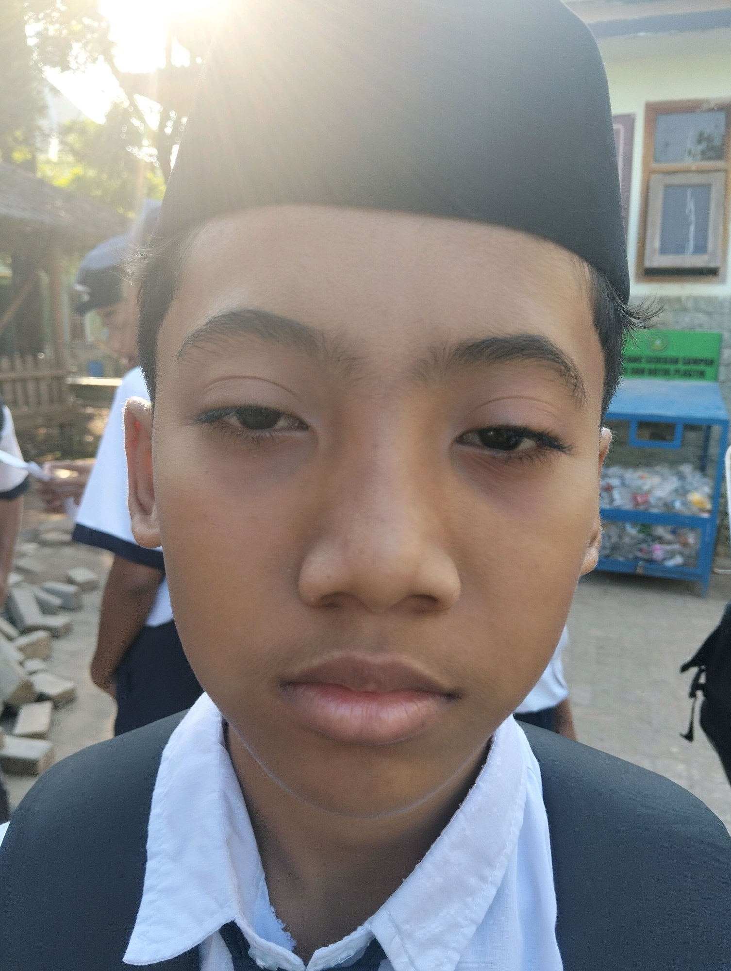 A medium shot, eye-level outdoor photograph captures the front-facing head and shoulders of a young male wearing a black cap and a white collared shirt. The subject's face, illuminated by bright sunlight from the upper left, is the primary focus. The background shows a building with windows and a blue recycling bin filled with plastic bottles. In the distance, other individuals and a wooden fence are visible, suggesting a public or educational setting. The image appears to have been taken in Paciran, Indonesia, as indicated by the context.